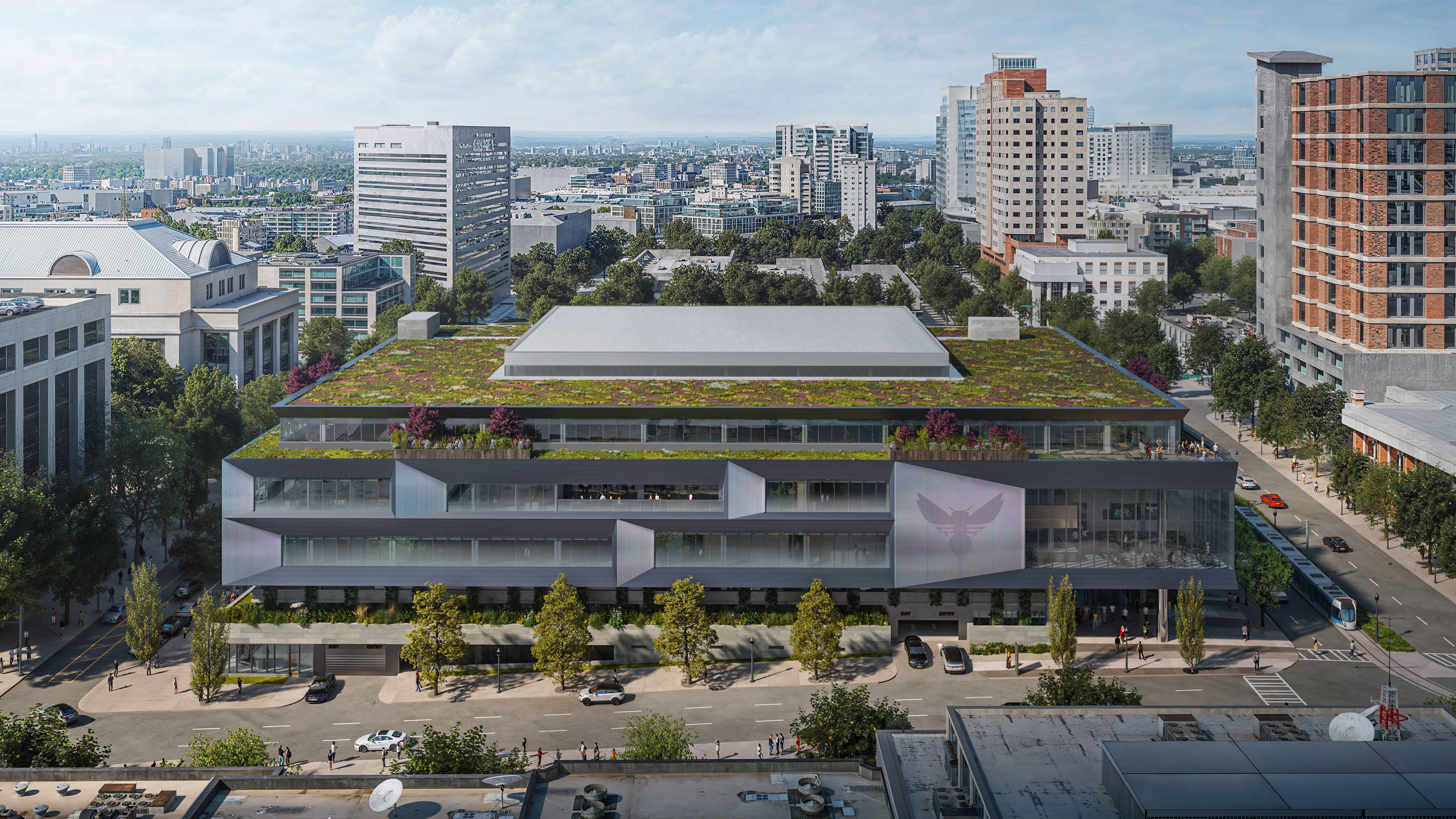 Aerial view of a city center: a large modern building with a lush green rooftop garden in the foreground, surrounded by trees, streets, and numerous high-rise buildings and a distant skyline.