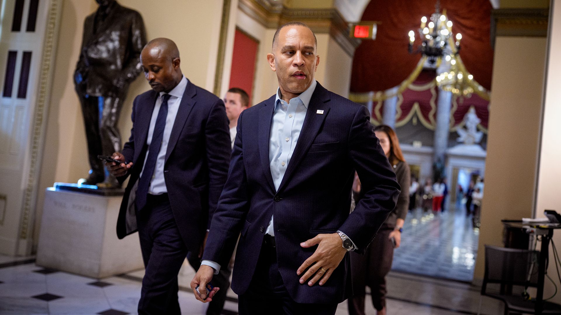 House Minority Leader Hakeem Jeffries, wearing a blue suit with no tie, walks through the ornate halls of the U.S. Capitol flanked by a coterie of aides.