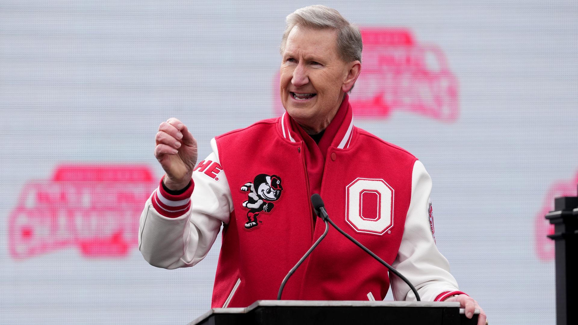 Ohio State president Ted Carter speaks at a podium, wearing a scarlet-and-white varsity jacket with a large block O patch.