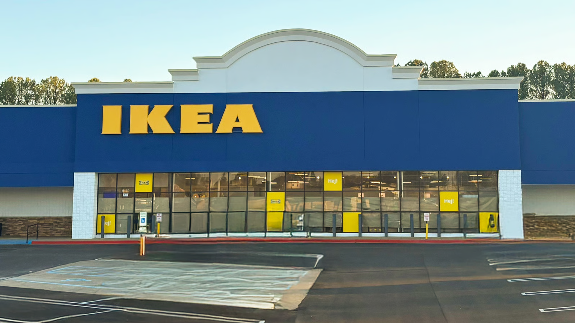 Front view of an IKEA store with blue and white exterior, large yellow IKEA sign, glass windows, and empty asphalt parking lot in front during daytime.