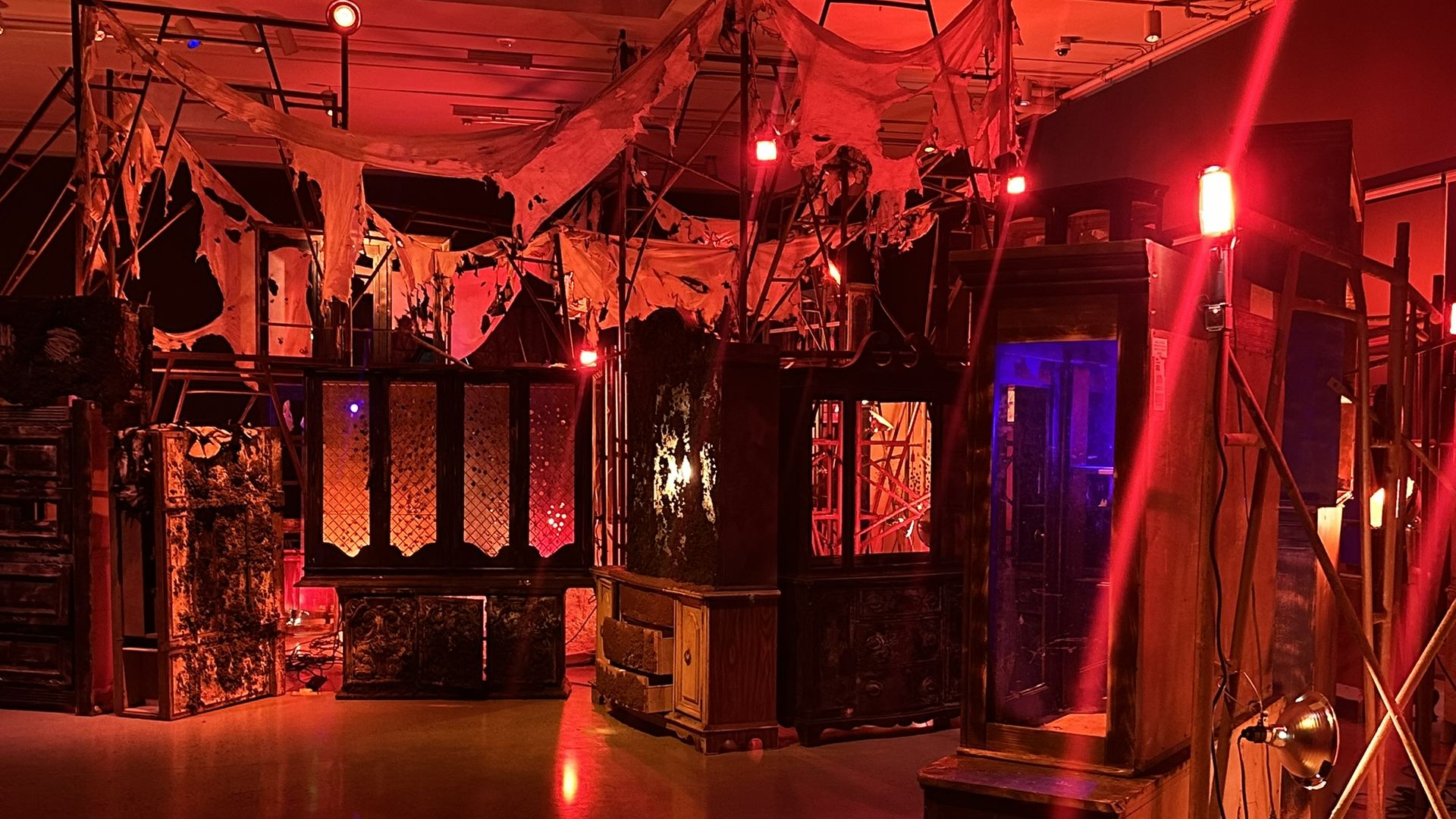 Inside a theater stage bathed in red light, torn fabric hangs from metal scaffolding and wooden structures. Cables and props litter the scene, giving a spooky, abandoned look.