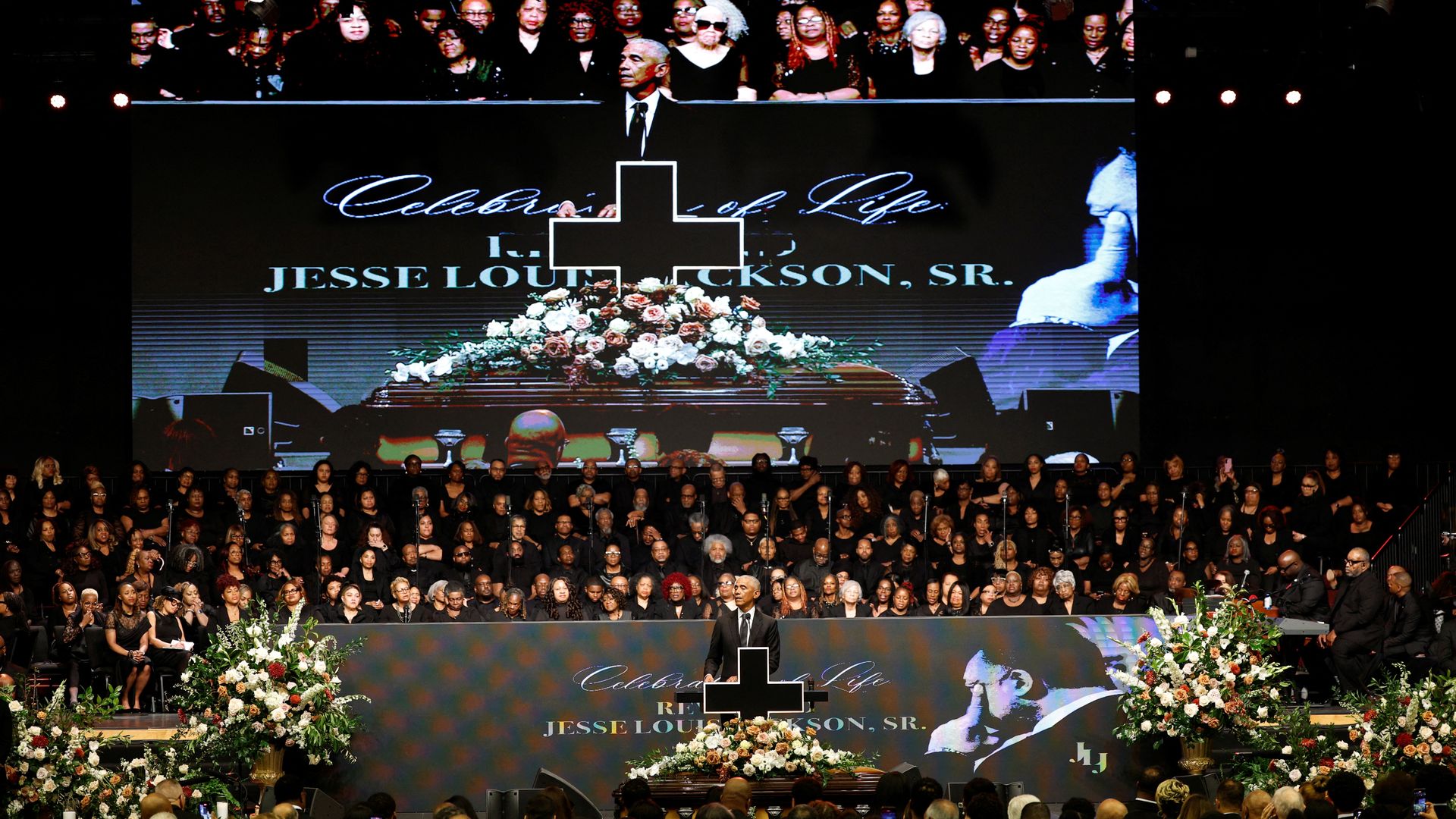 Large indoor funeral service for Jesse Louis Jackson Sr. with speaker at podium, choir dressed in black, a decorated casket, floral arrangements, and a large screen displaying the event.