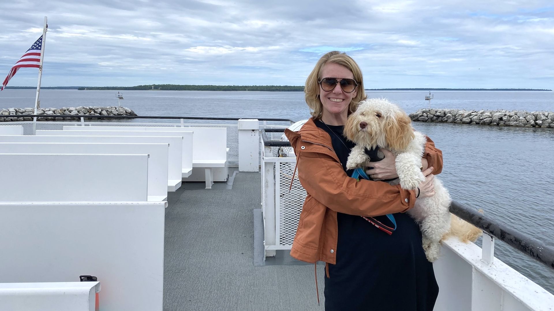 A woman in an orange jacket holding a white shaggy dog on a ferry.