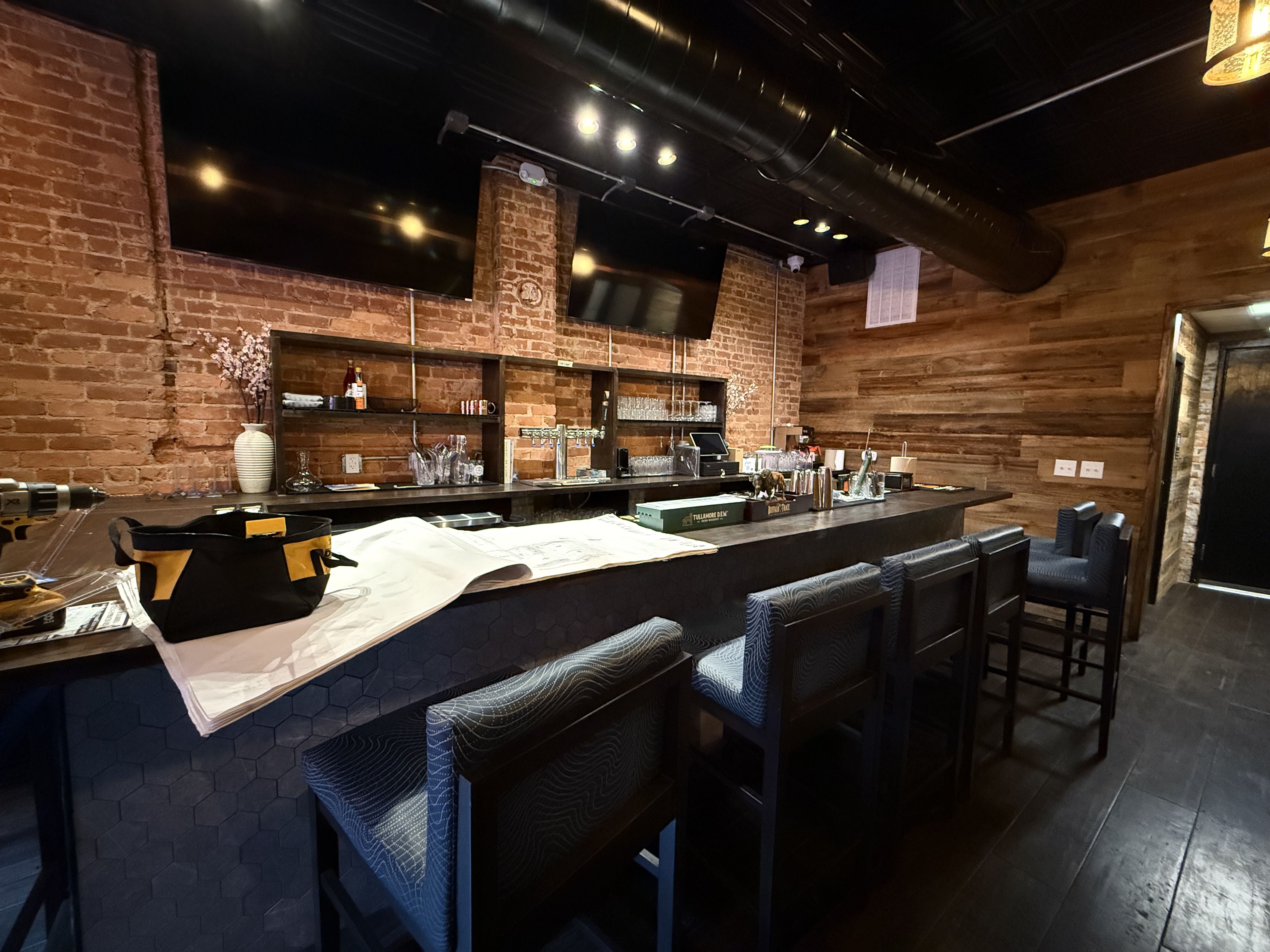 Interior of a bar with exposed brick and wood walls, dark hexagonal counter, padded bar stools, two large TVs, drink glasses, and a work bag with plans on the counter.