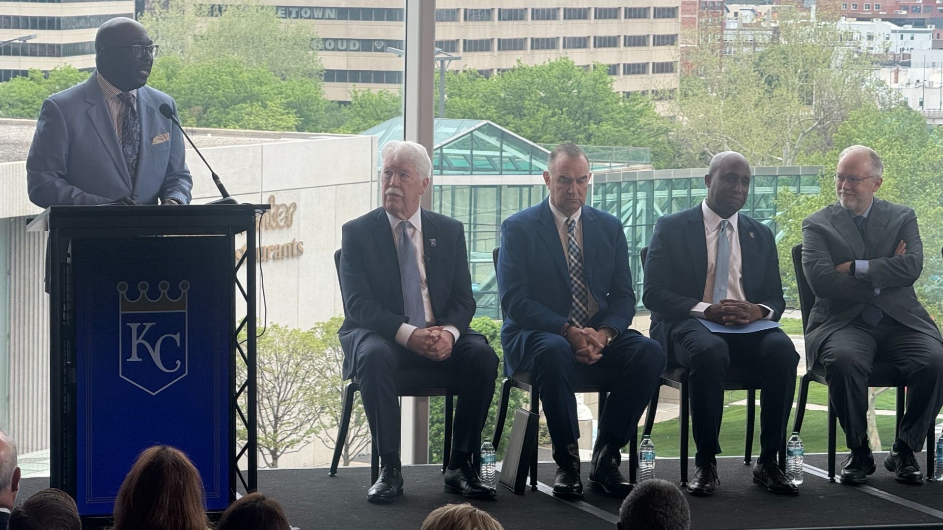A man in a blue suit speaks at a podium with a KC crest; four suited men sit on a panel outdoors, with a glass structure and green trees behind them.