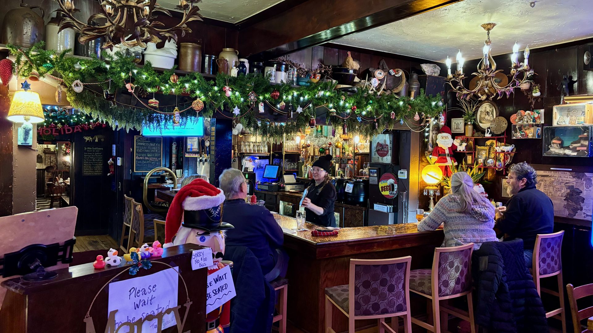 Cozy pub interior with wooden bar, garland and ornaments hanging above, chandeliers lit, a nutcracker statue with a Santa hat, and patrons sitting and talking with a bartender.