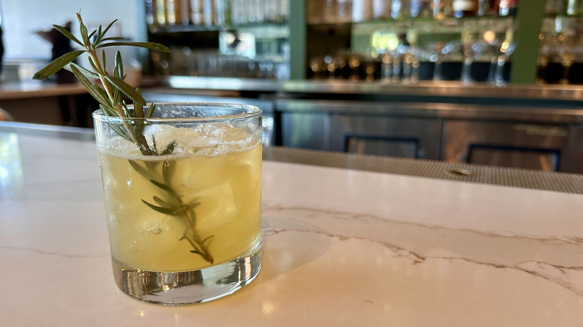 A photo of a yellow-ish cocktail with a rosemary garnish on a bar top with bottles of liquor on a shelf in the background. 