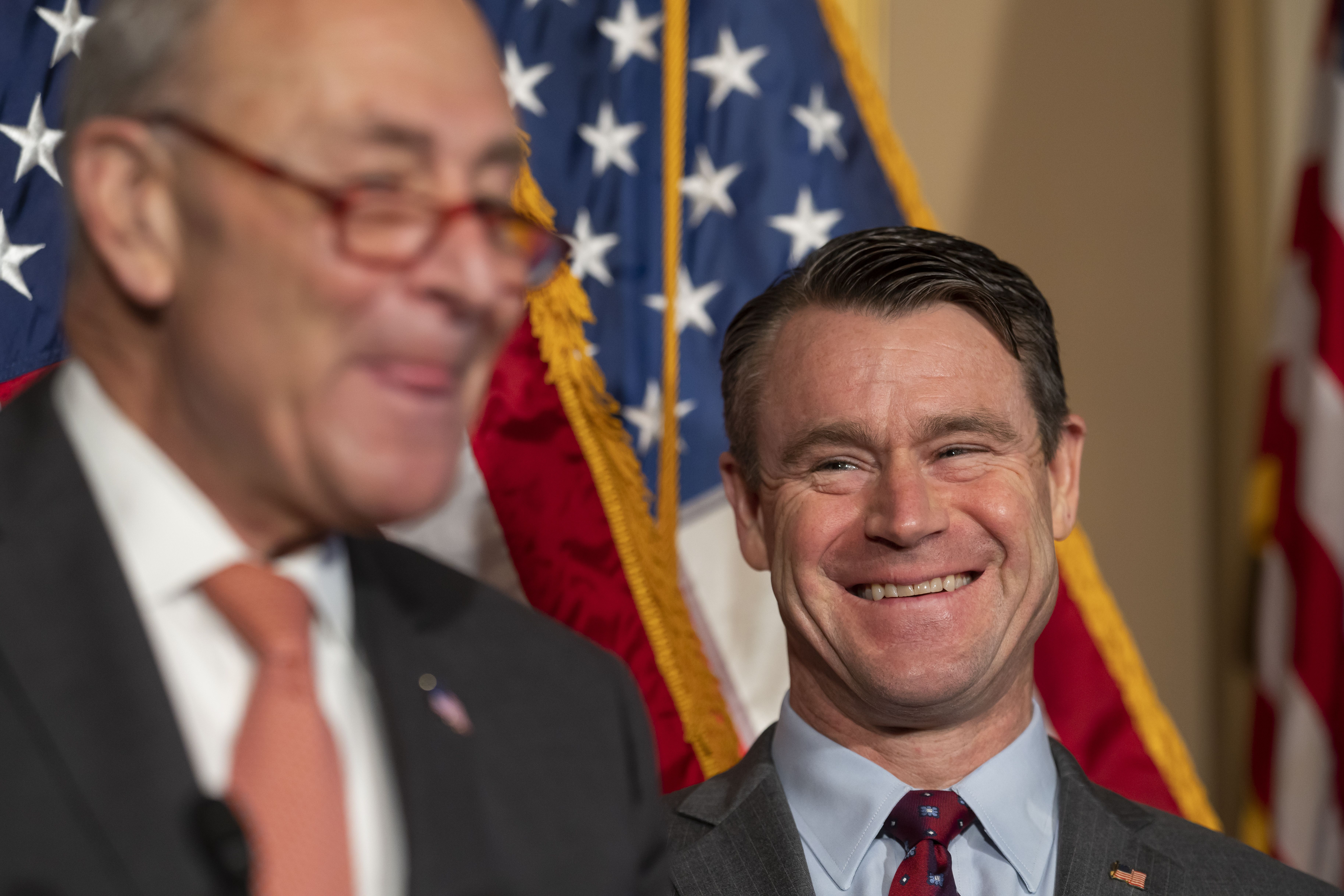 Sen. Todd Young smiles behind Senate Majority Leader Chuck Schumer at a press conference.