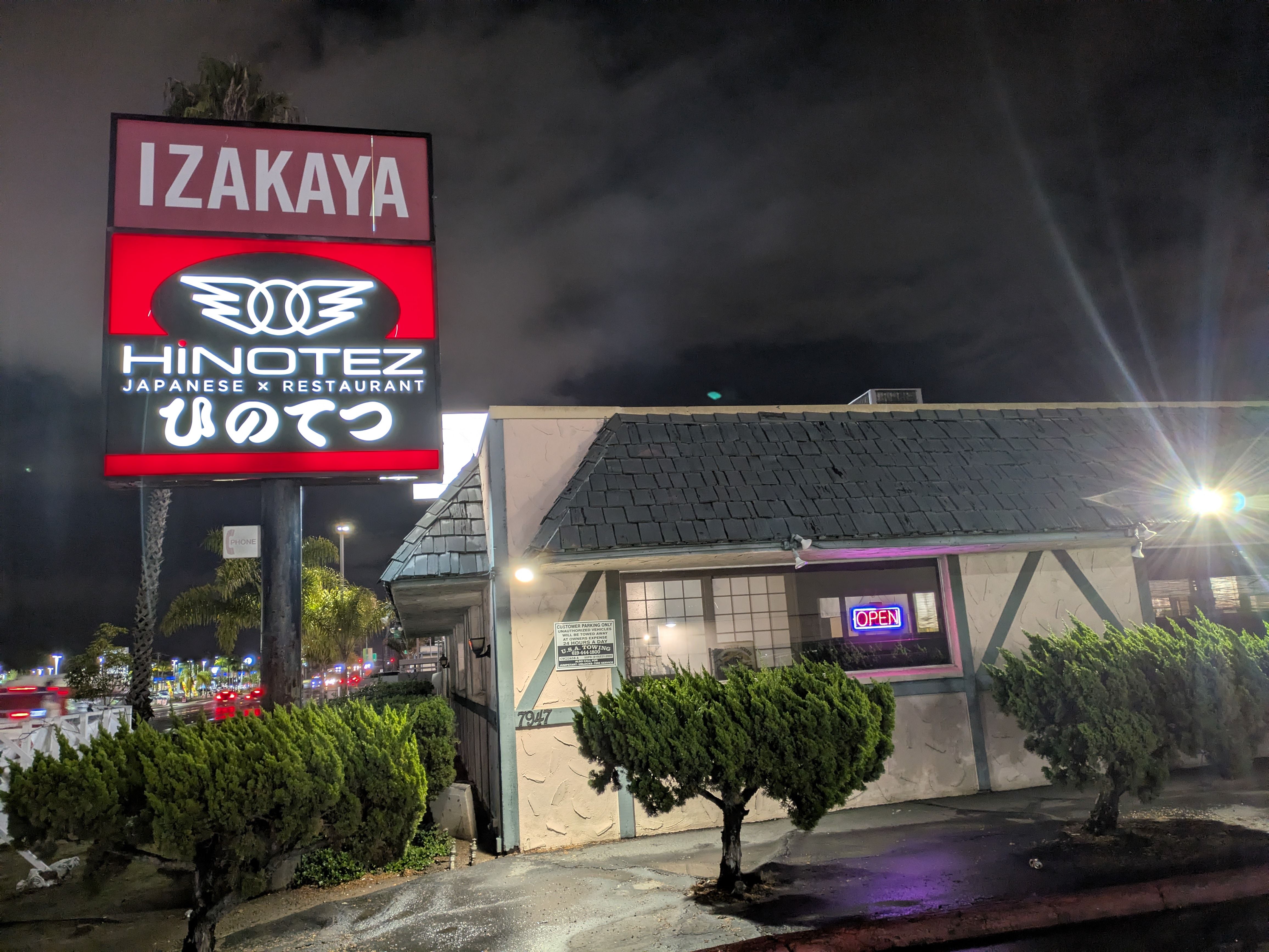 Night view of a Japanese restaurant named HiNotez with a red and white illuminated sign reading IZAKAYA and an open neon sign in the window of the small building.