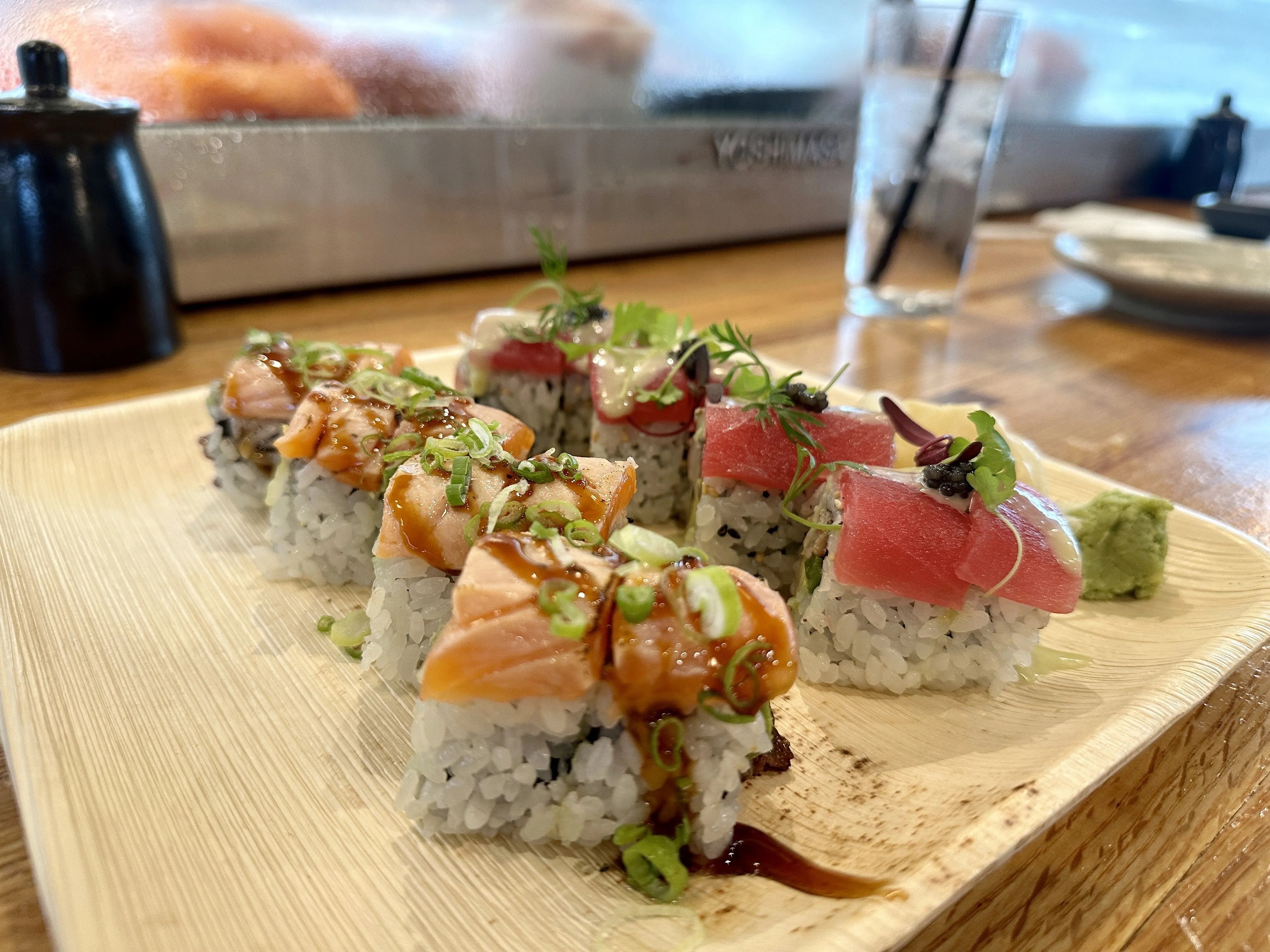 Plate with sushi rolls topped with orange salmon, red tuna, green onions, microgreens, and sauce, served with a dollop of green wasabi on a wooden table with a glass of water in the background.