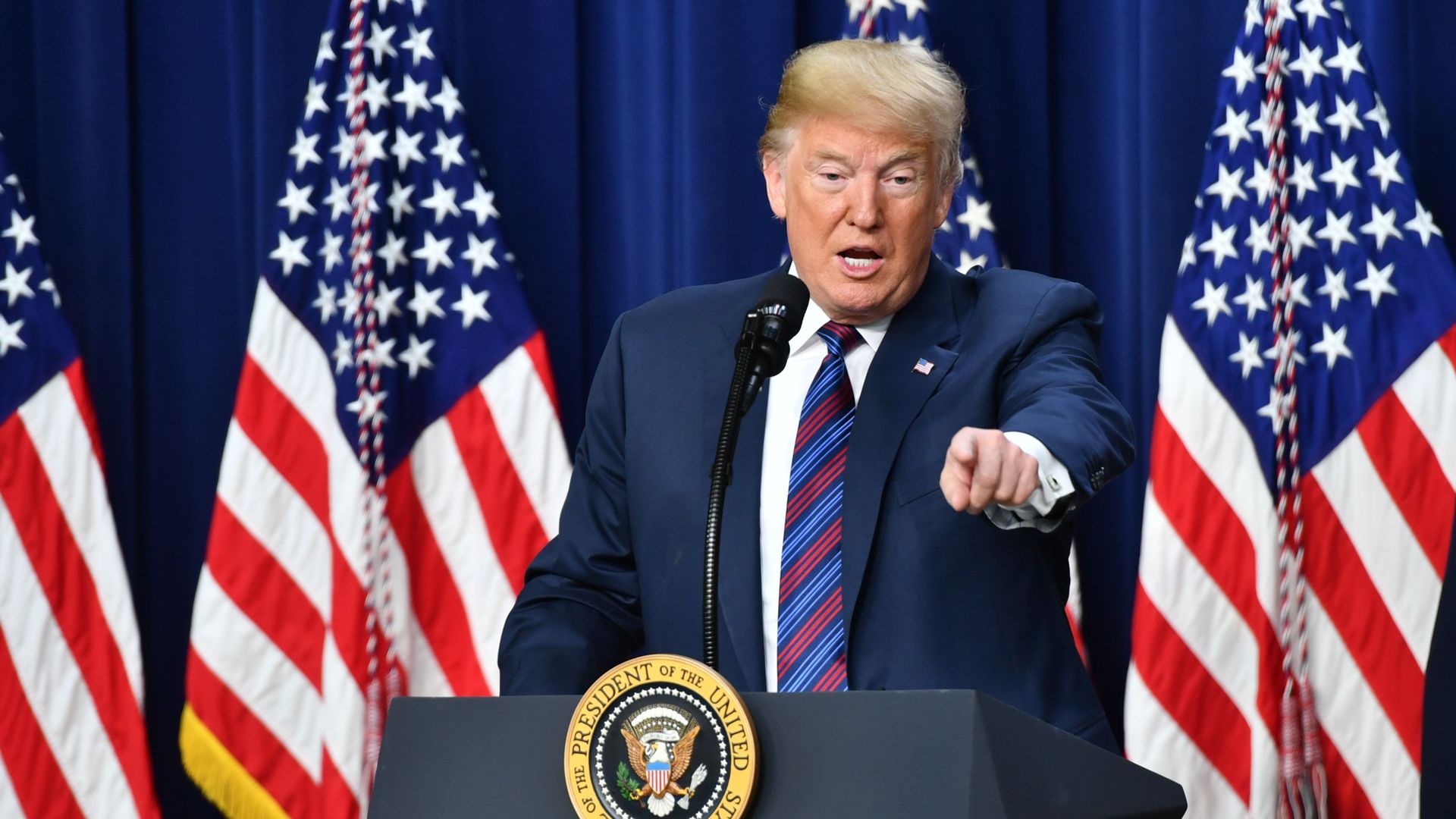 Trump stands before American flags before a blue drape, and points.
