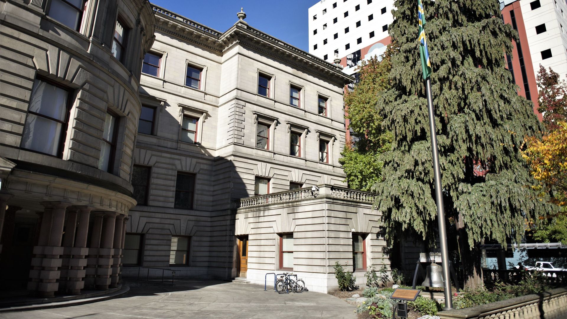 Historic stone building with rounded corner and many windows under clear blue sky. Tall evergreen tree and flagpole with green flag near building entrance. A bicycle parked nearby.