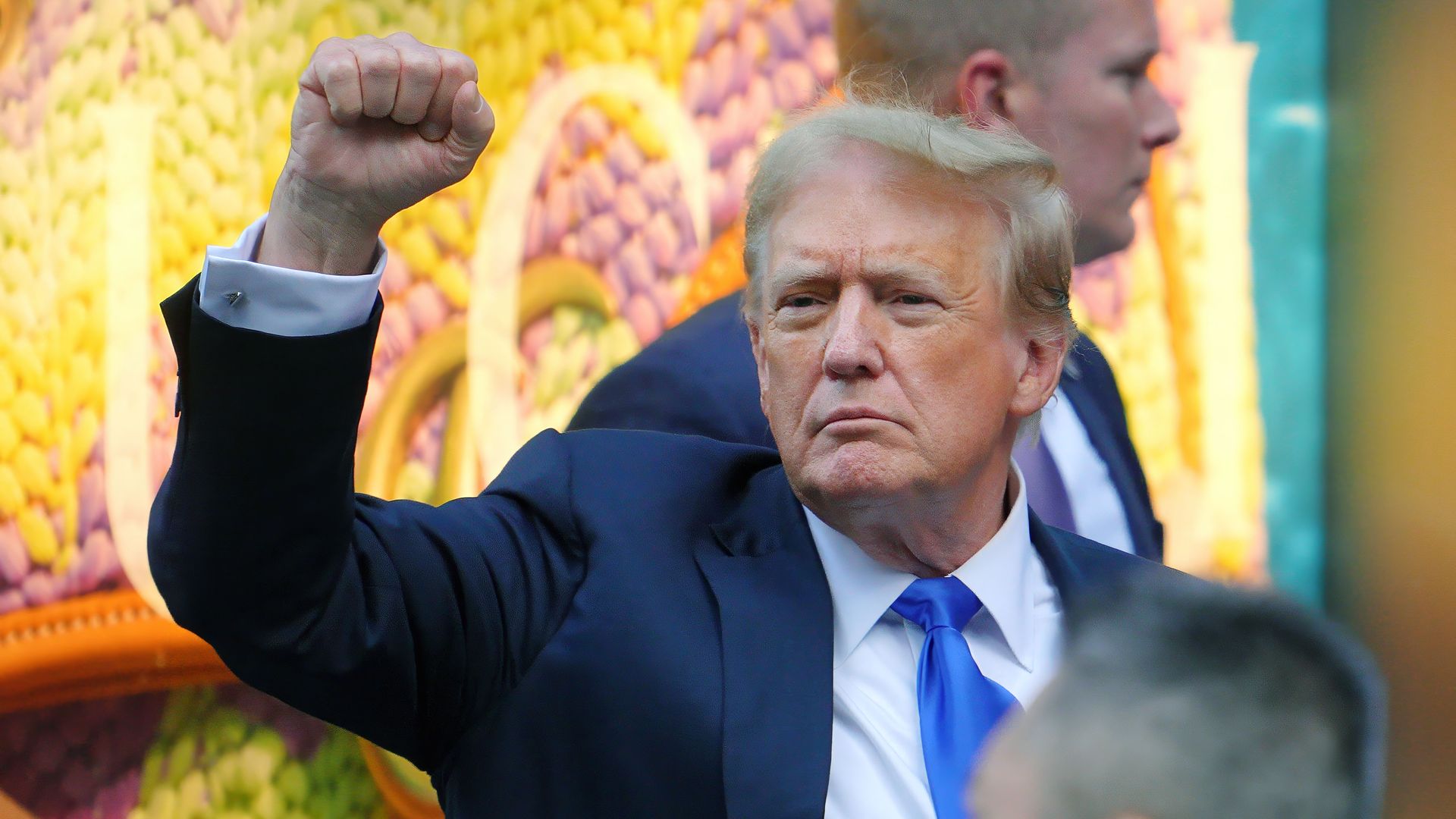 Former President Trump, wearing a blue suit and holding his fist in the air in New York City.