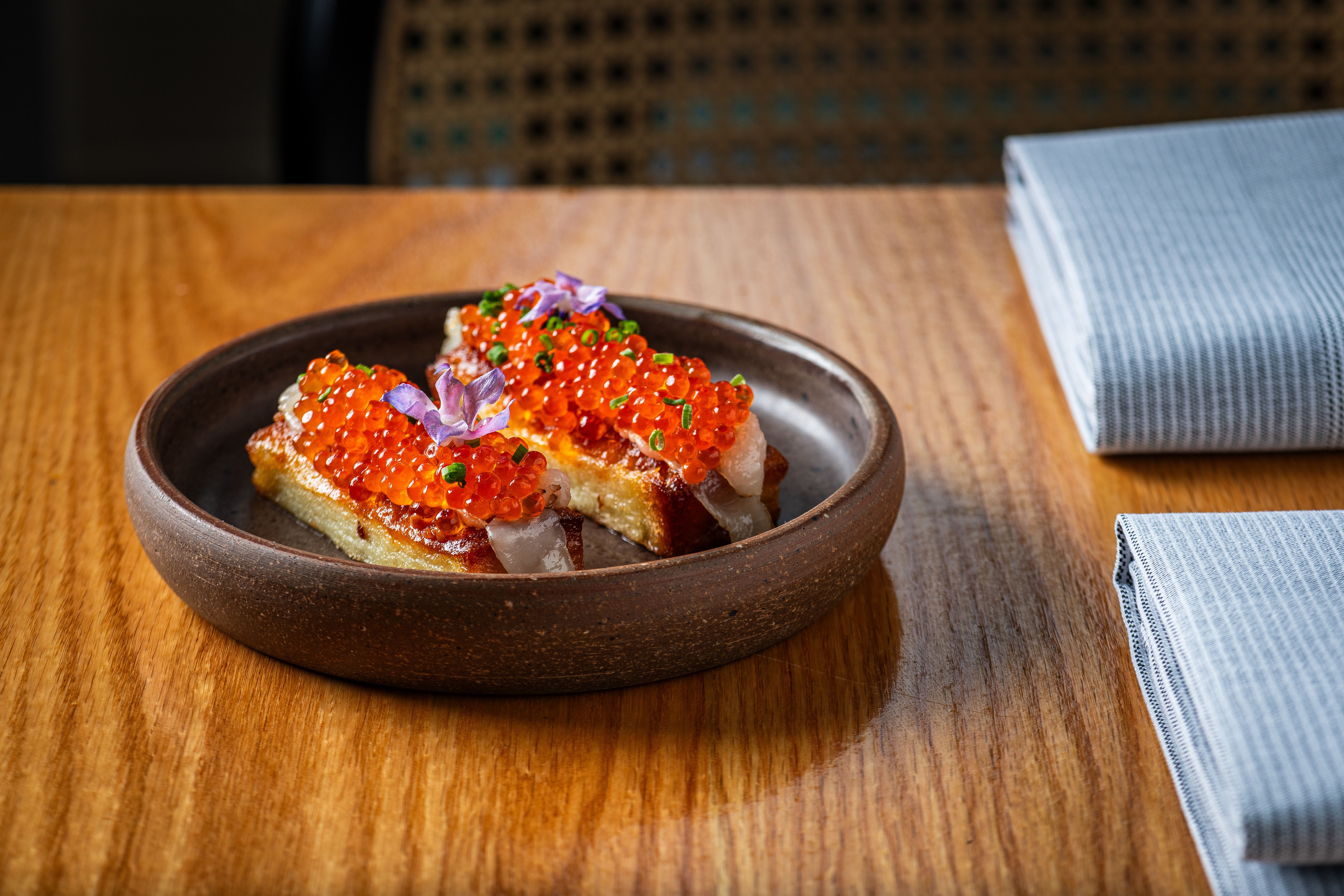 Two rectangular pieces of cake topped with red-orange fish roe, a purple edible flower, and green garnish, served on a round brown dish on a wooden table with folded blue striped napkins nearby.