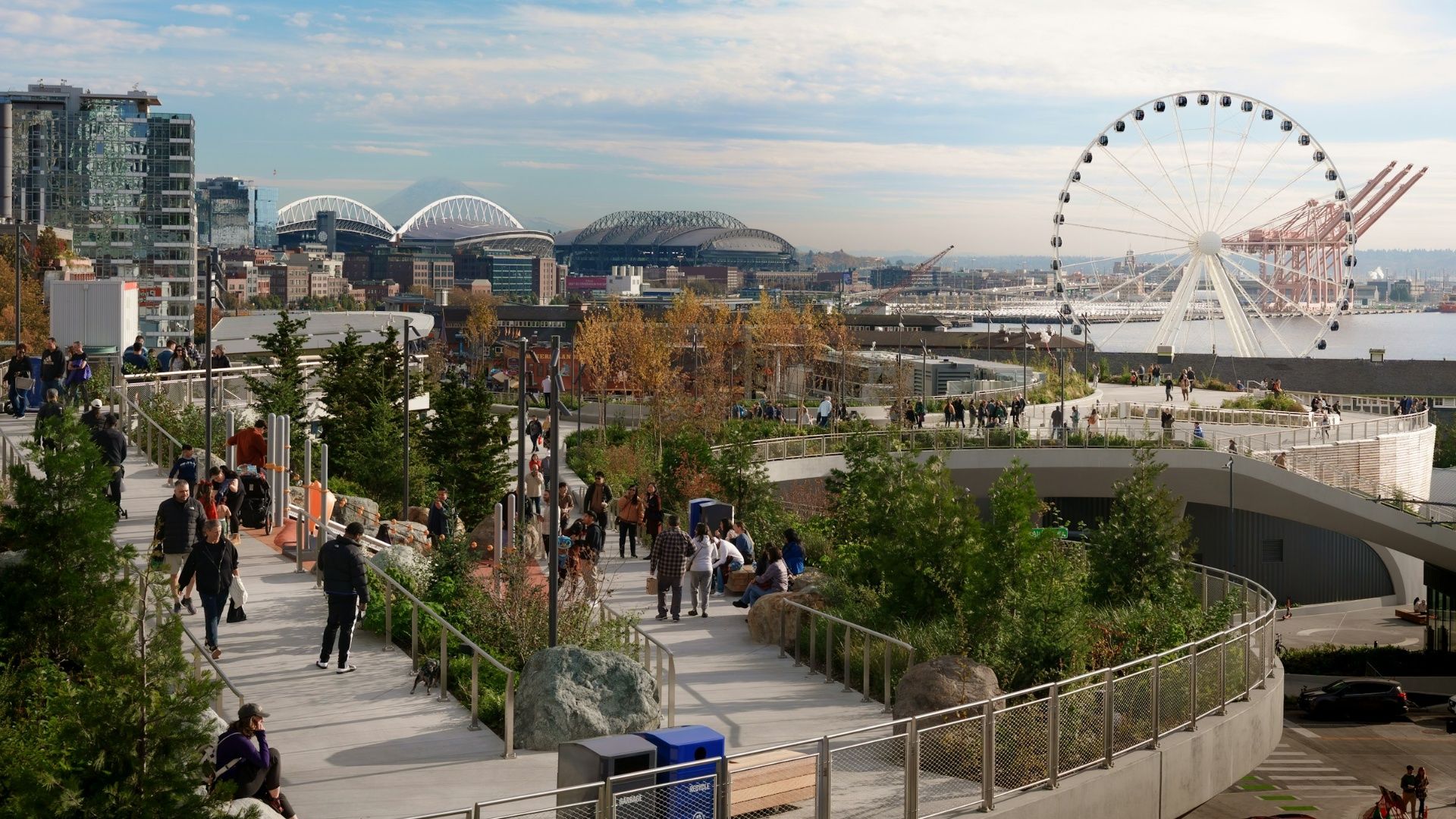 Wide city waterfront with a large white Ferris wheel by the bay, modern glass buildings, arched-roof stadiums, cranes, and a busy elevated promenade lined with trees and pedestrians.