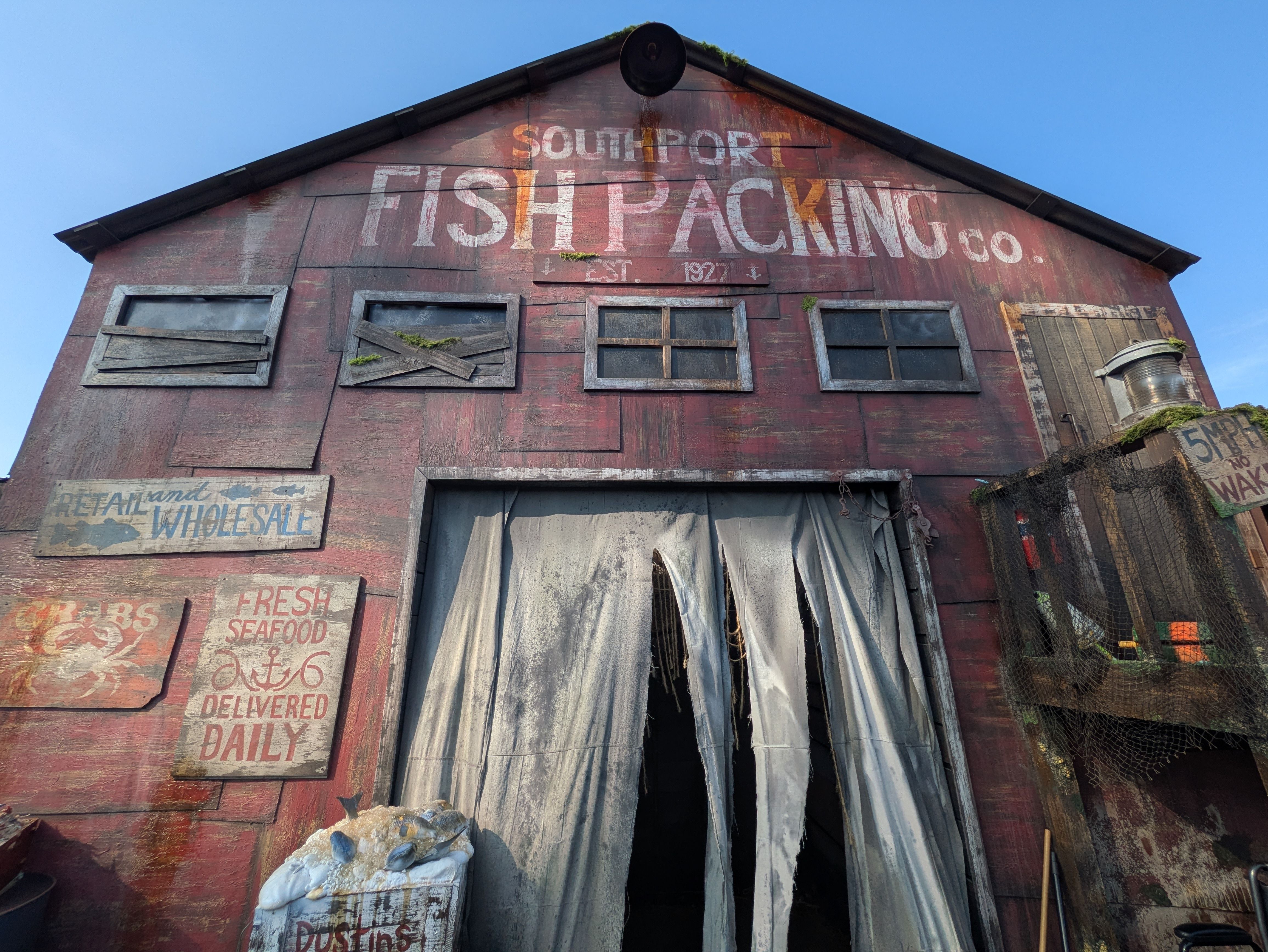 Old red wooden fish-packing warehouse with a faded sign reading "Southport Fish Packing Co.", moss on the roof, boarded windows, torn curtains at the entrance, and a clear blue sky.