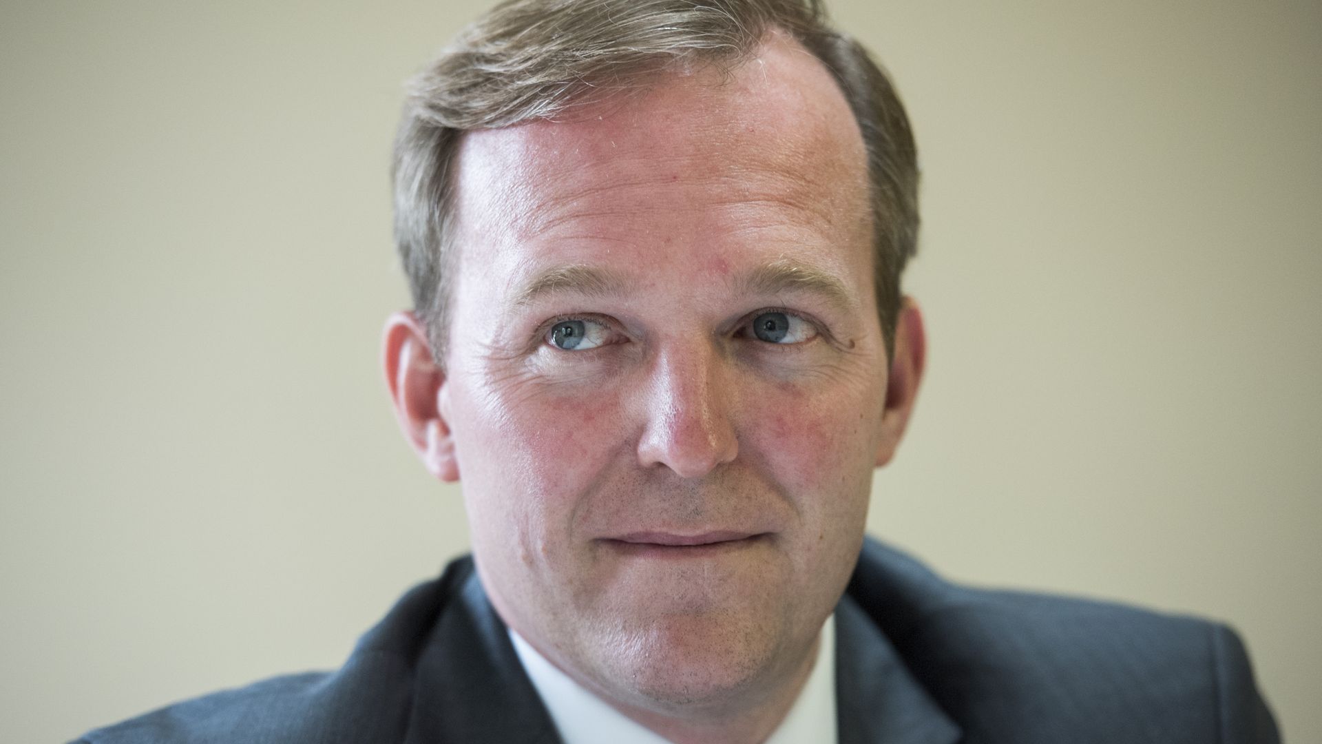Close-up of a man in a dark suit and striped tie, looking slightly off-camera. He has light brown hair, blue eyes, and a pale beige background.