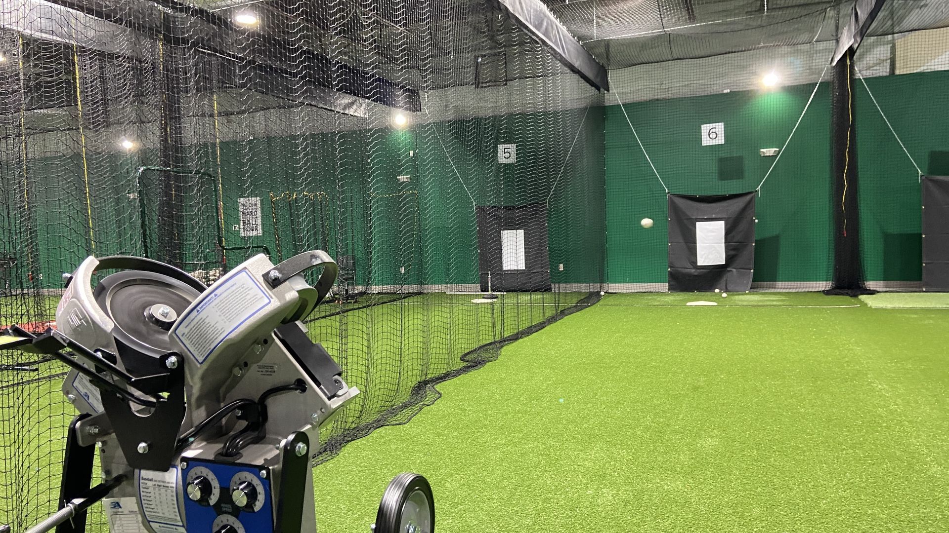 A baseball soars through the air from home plate out of a machine at an indoor facility.