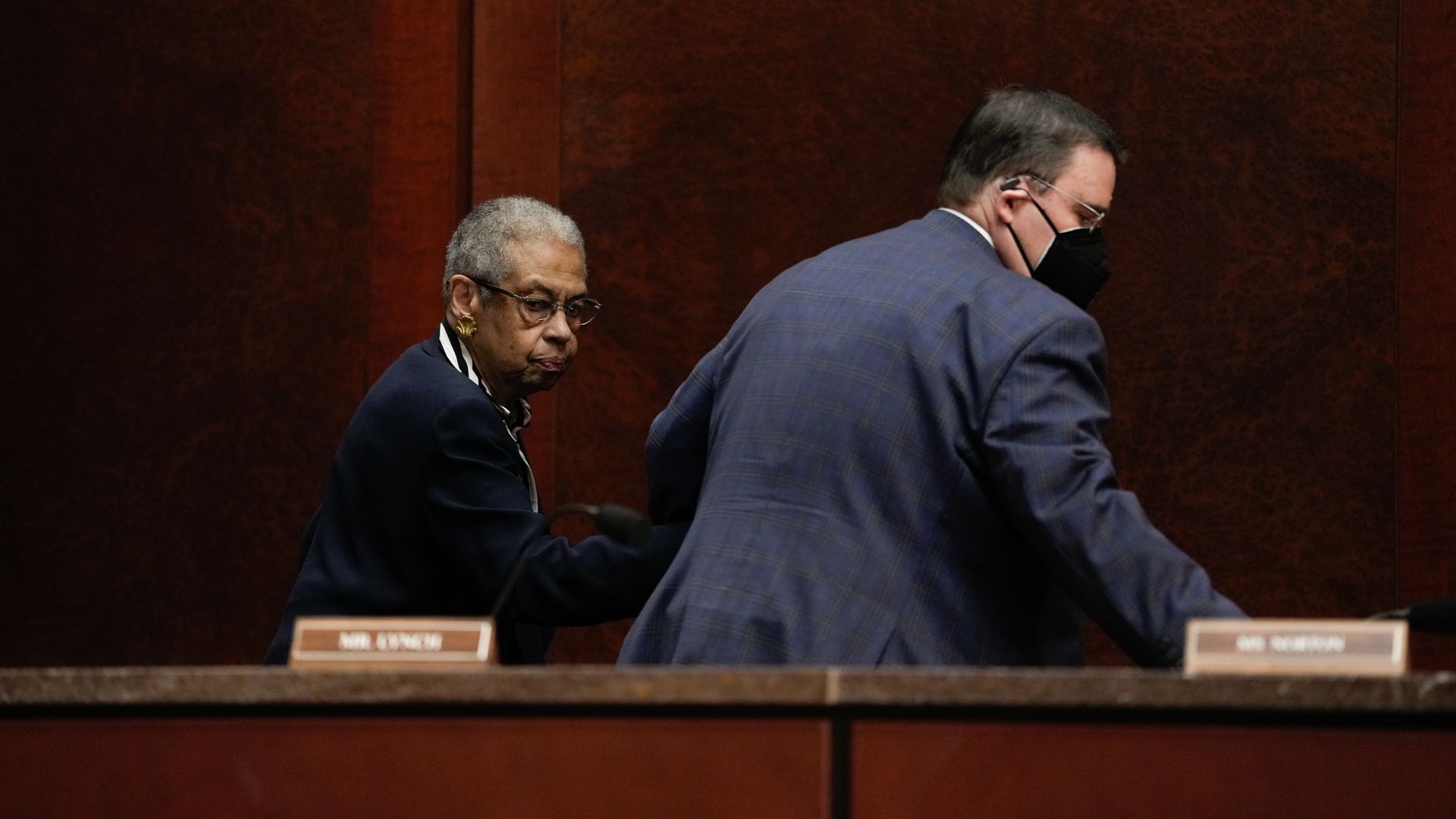 A woman in a dark outfit and glasses looks back while a man in a blue jacket and black face mask moves away in a wood-paneled room with nameplates on the desk.