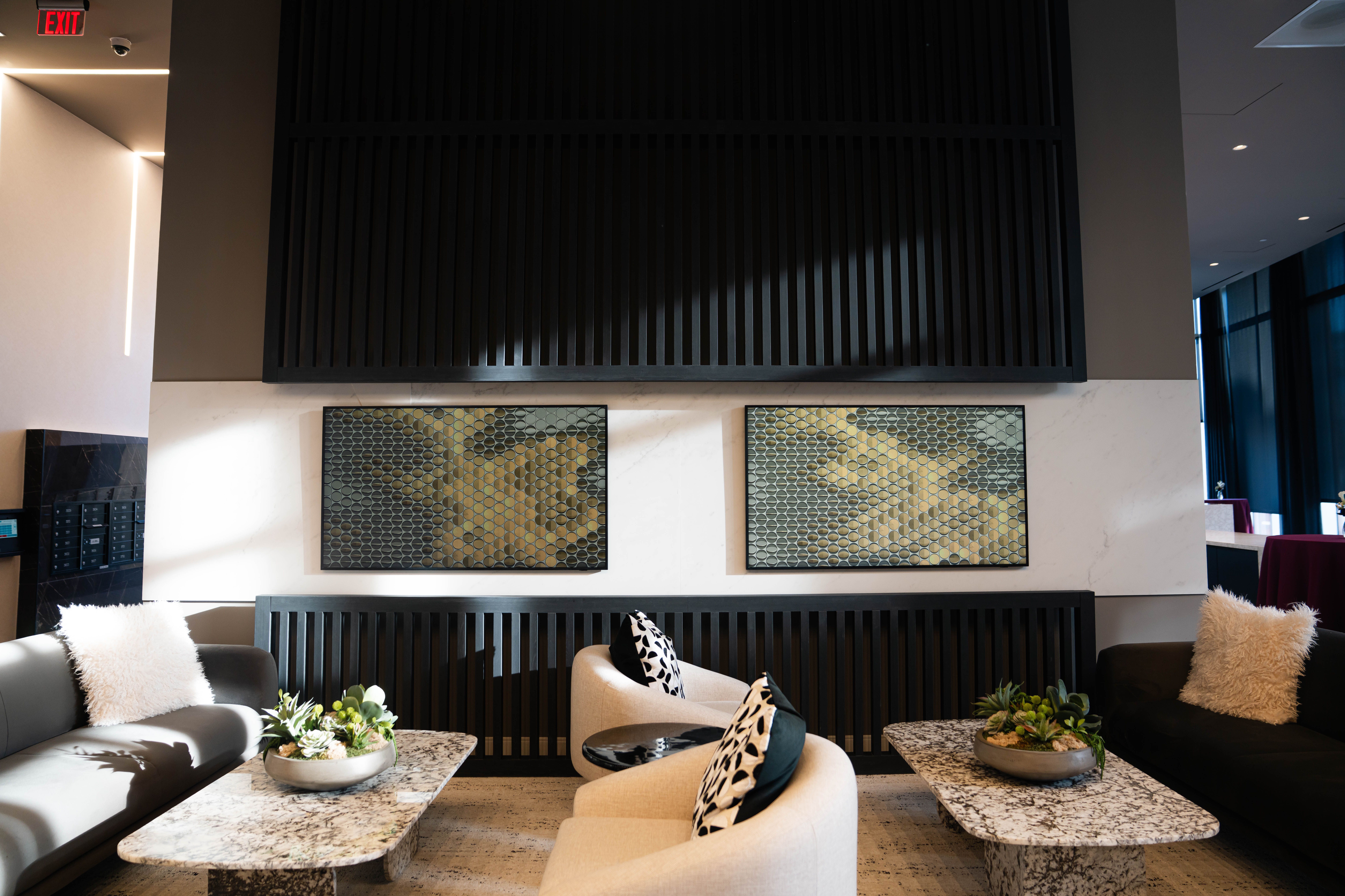Modern seating area with two beige chairs and two dark sofas, each sofa has a white fluffy pillow. Two marble coffee tables feature small green succulent arrangements. Dark wall panels and hexagonal art.