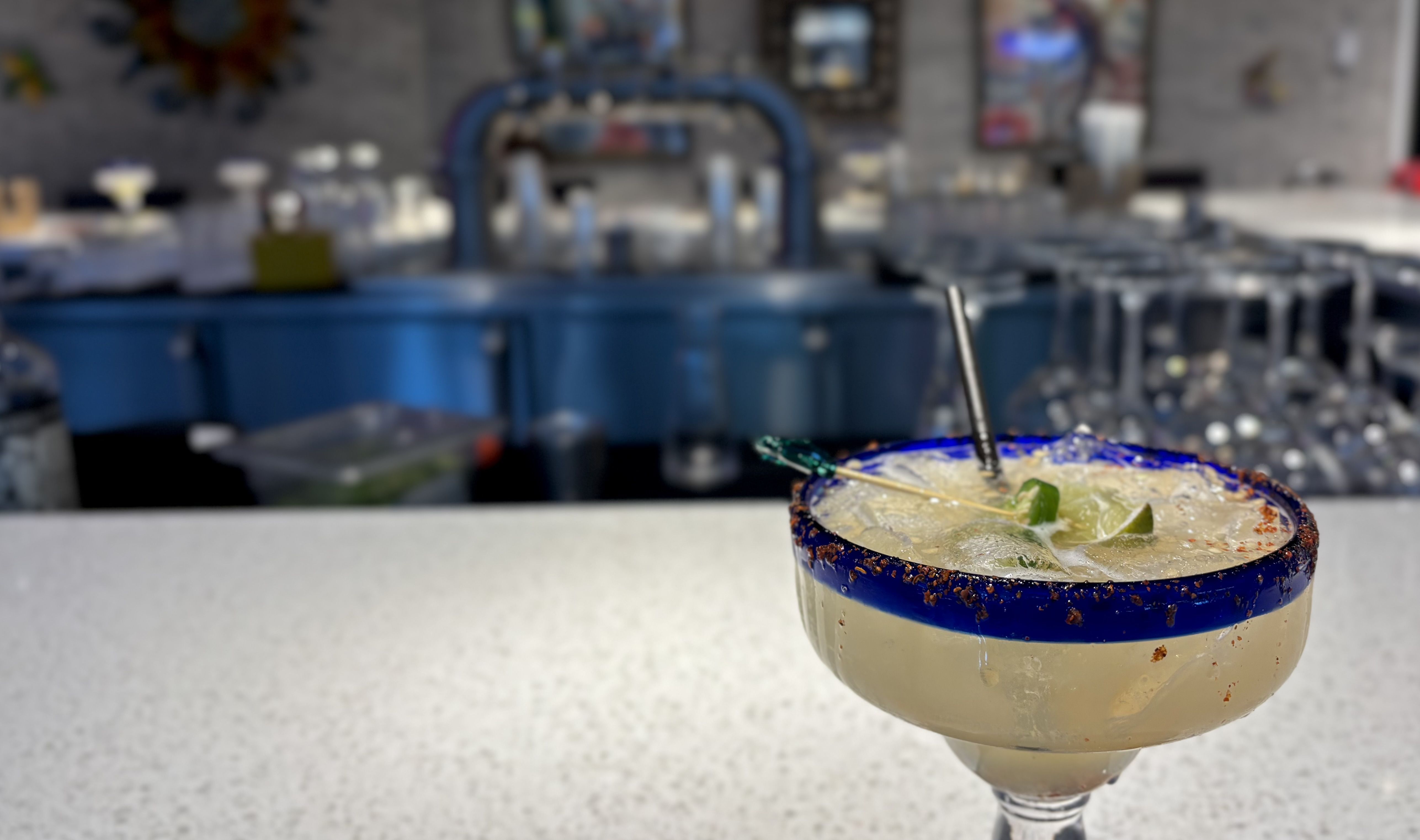 A close-up of a salted margarita in a blue-rimmed stem glass with a lime wedge and straw, set against a blurred bar or kitchen backdrop.