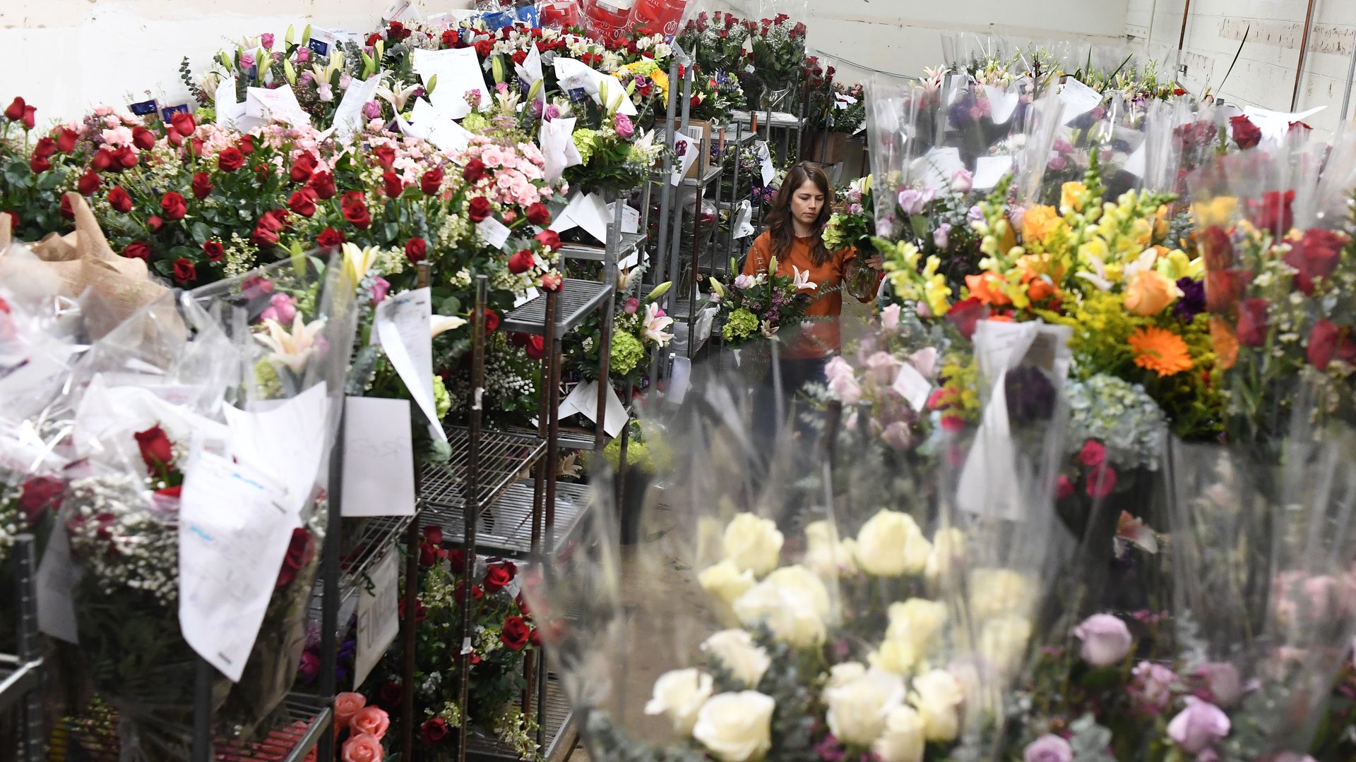 Various flowers and bouquets are placed near one another, with several sitting on shelves with multiple cases. In the middle background, a woman is seen handling two small bouquets. 