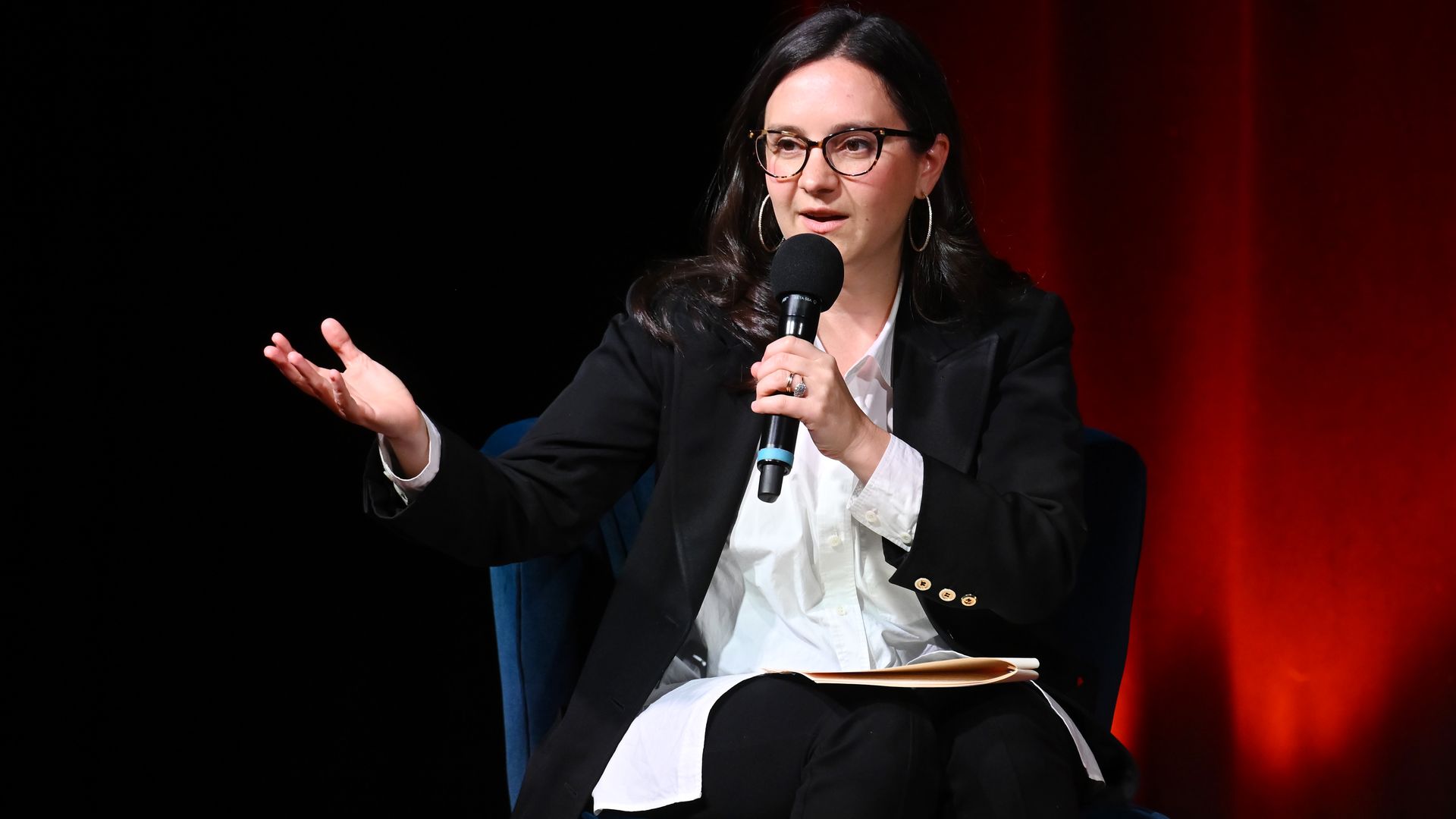 Woman with glasses and hoop earrings sitting on a blue chair, speaking into a microphone, wearing a black blazer and white shirt, red curtain background with soft lighting