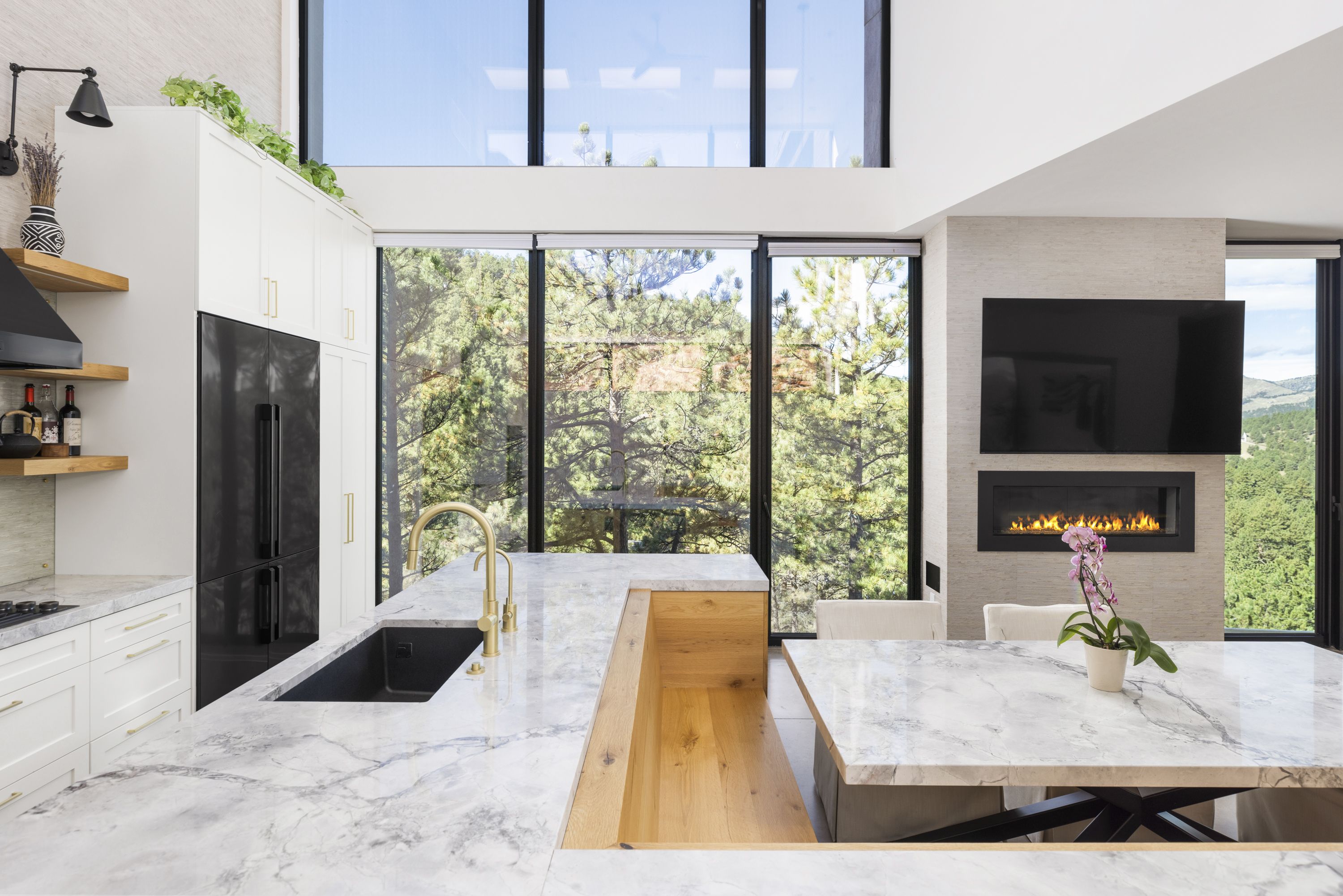 Modern kitchen with white cabinets, black refrigerator, marble countertops, gold faucet, large windows showing green trees, marble dining table with orchid, and wall-mounted TV above fireplace.