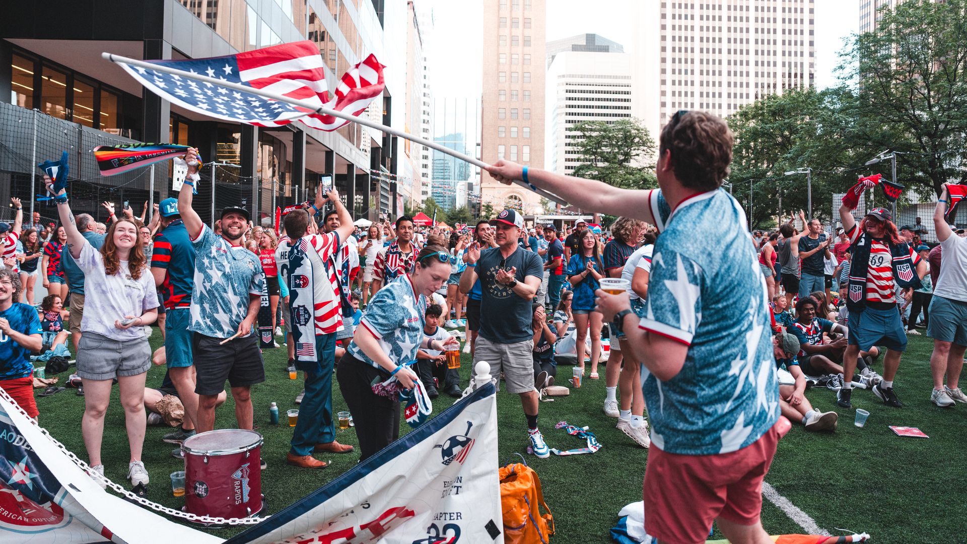 Crowded city parade scene with spectators in red, white, and blue as a large American flag is waved on a pole; tall buildings behind, cheering crowd and banners.
