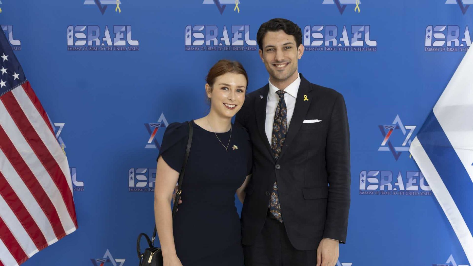 Sarah Lynn Milgrim stands next to Yaron Lischinsky as they pose for a photo. The backdrop reads "Israel" and they are flanked by American and Israeli flags. 