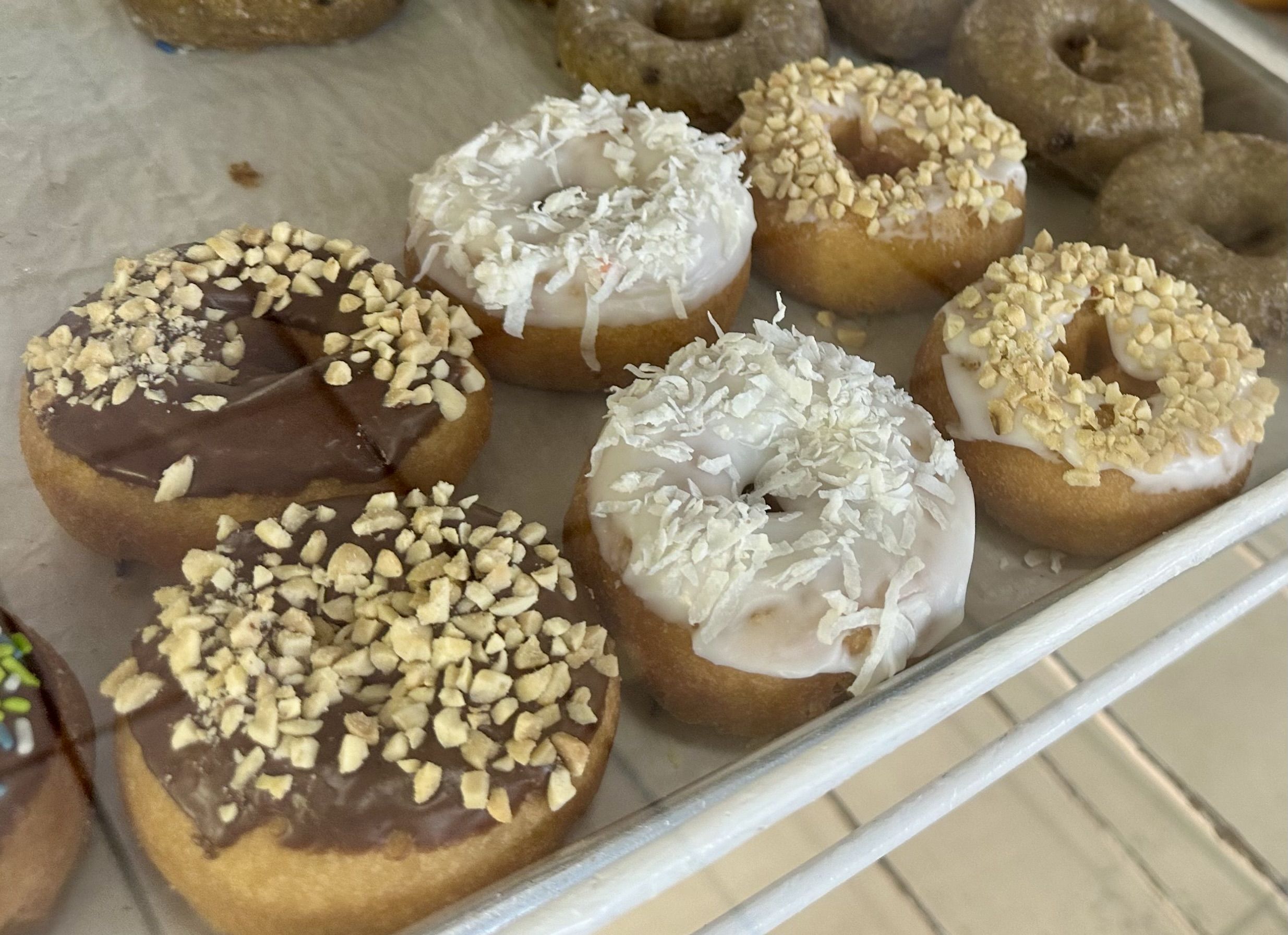Six doughnuts on a tray in a pastry case; two with nuts and chocolate frosting, two with white frosting and coconut, and another set with additional flavors.
