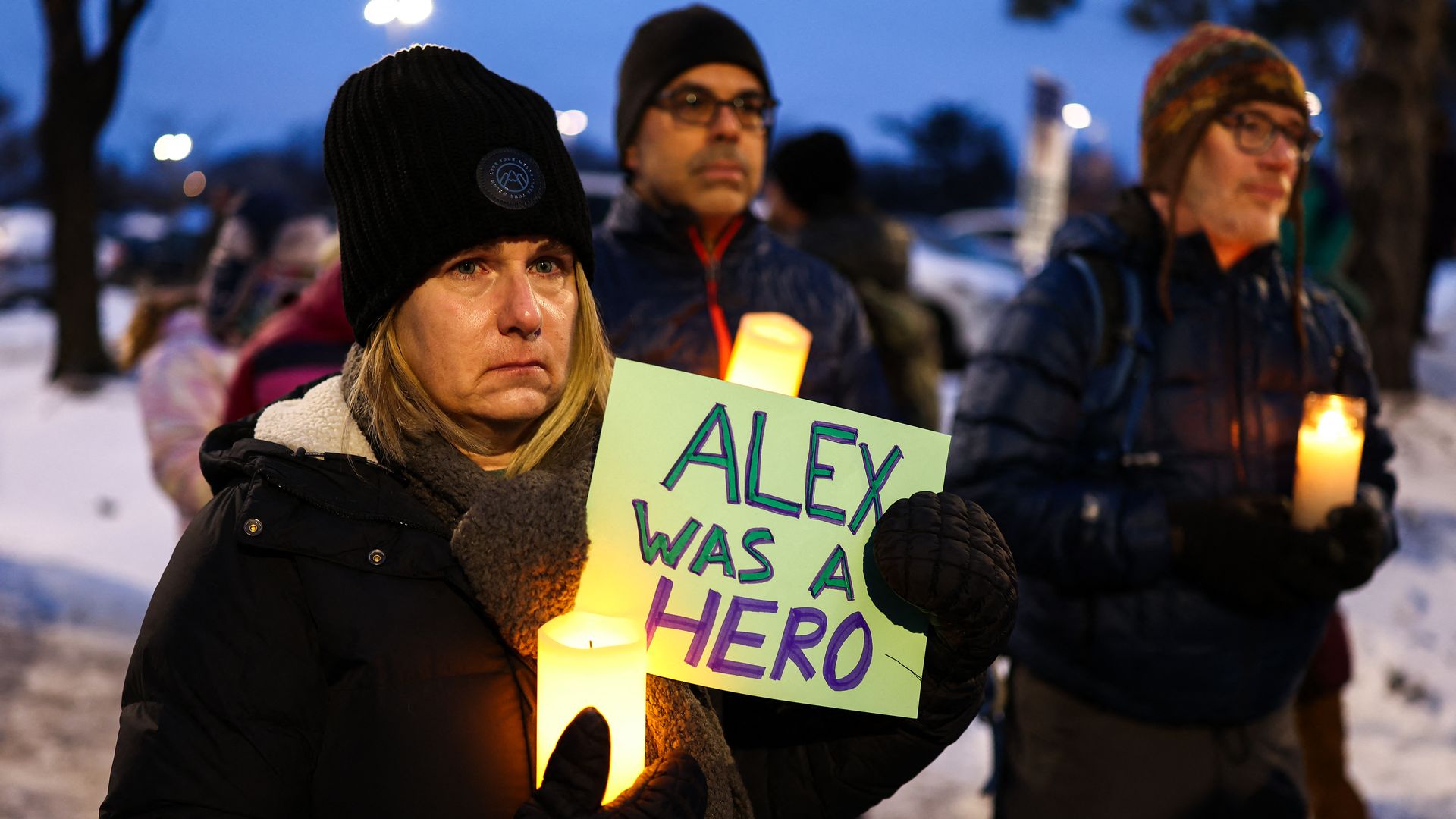 A group of people gather outside a memorial holding candles as one woman, wearing a black hat and winter coat holds a sign that reads "Alex was a hero" 