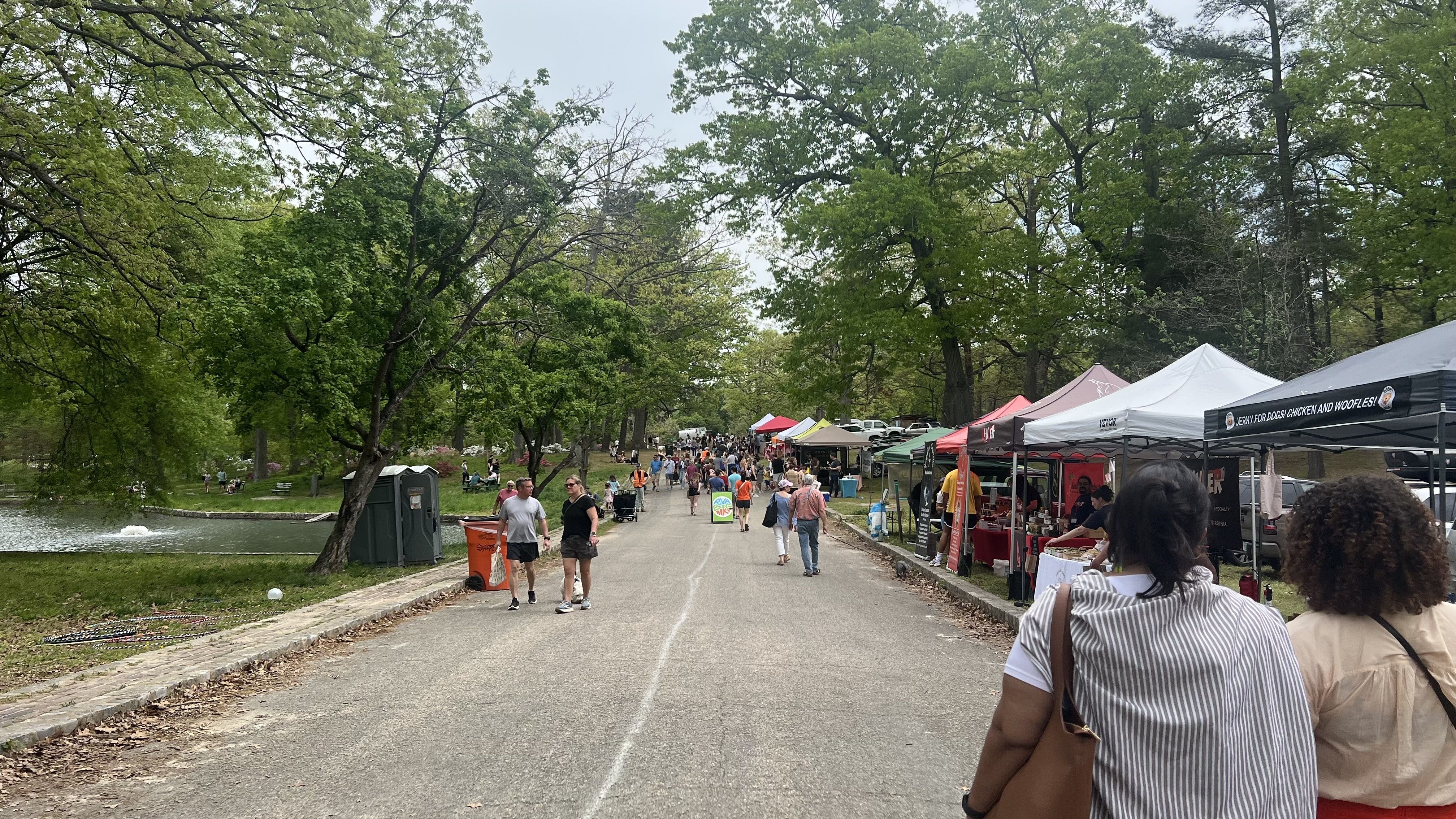 Tree-lined park path on a sunny day with colorful vendor tents on the right and people walking. A lake and green grass sit to the left as festival-goers browse stalls.