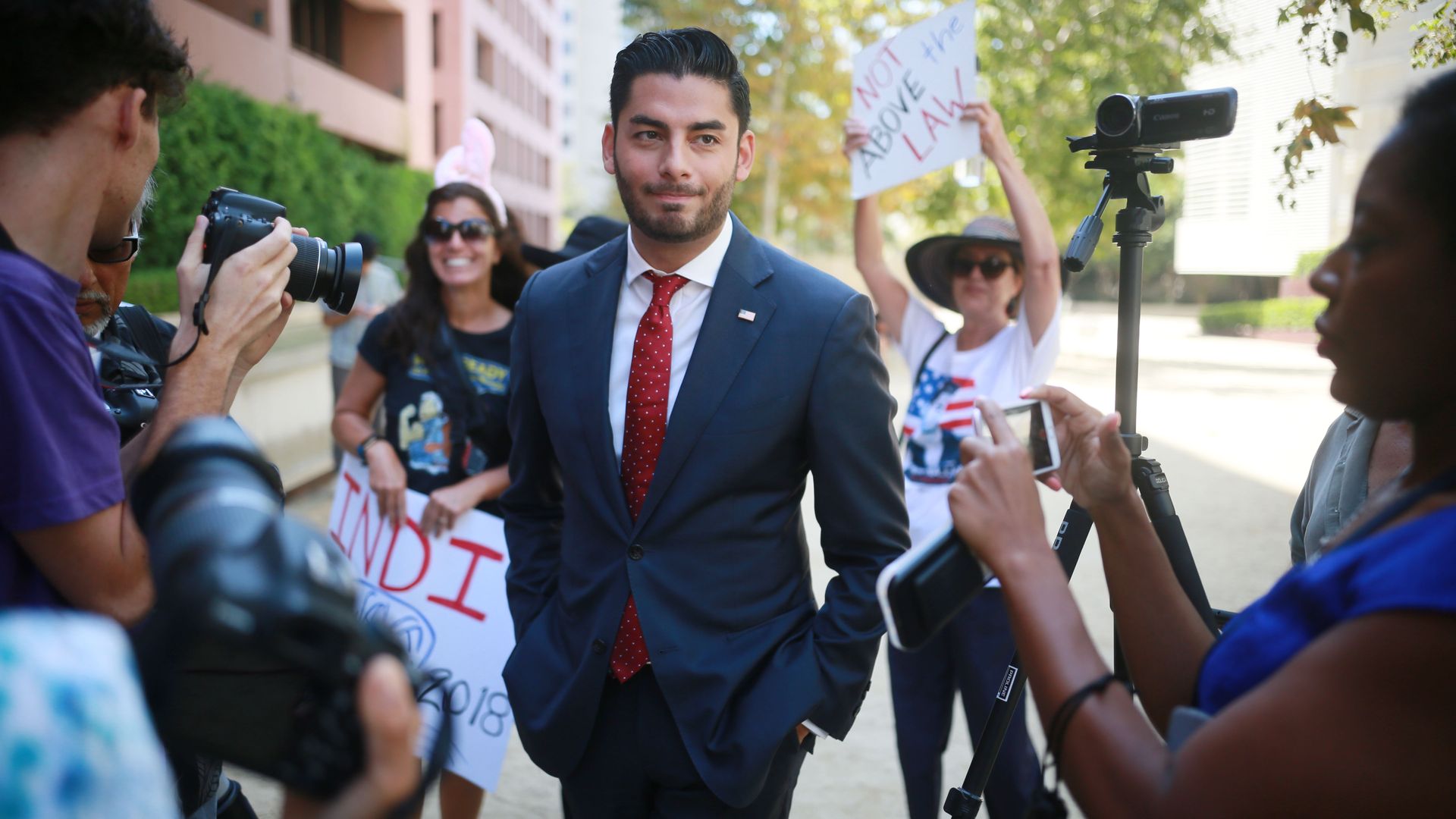 Man in a suit and red tie walks past photographers and protesters, one holding a sign