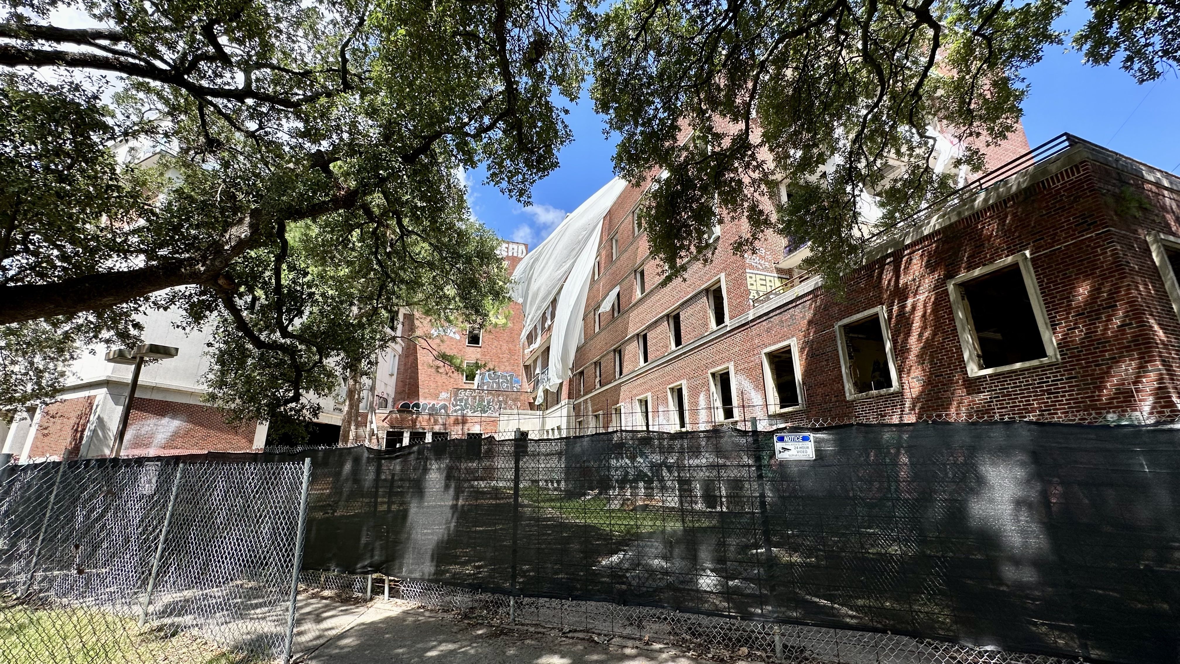 Abandoned red brick building with broken windows partially covered by white tarps, seen behind a chain-link fence. Large tree branches overhead, blue sky, graffiti on the lower wall.