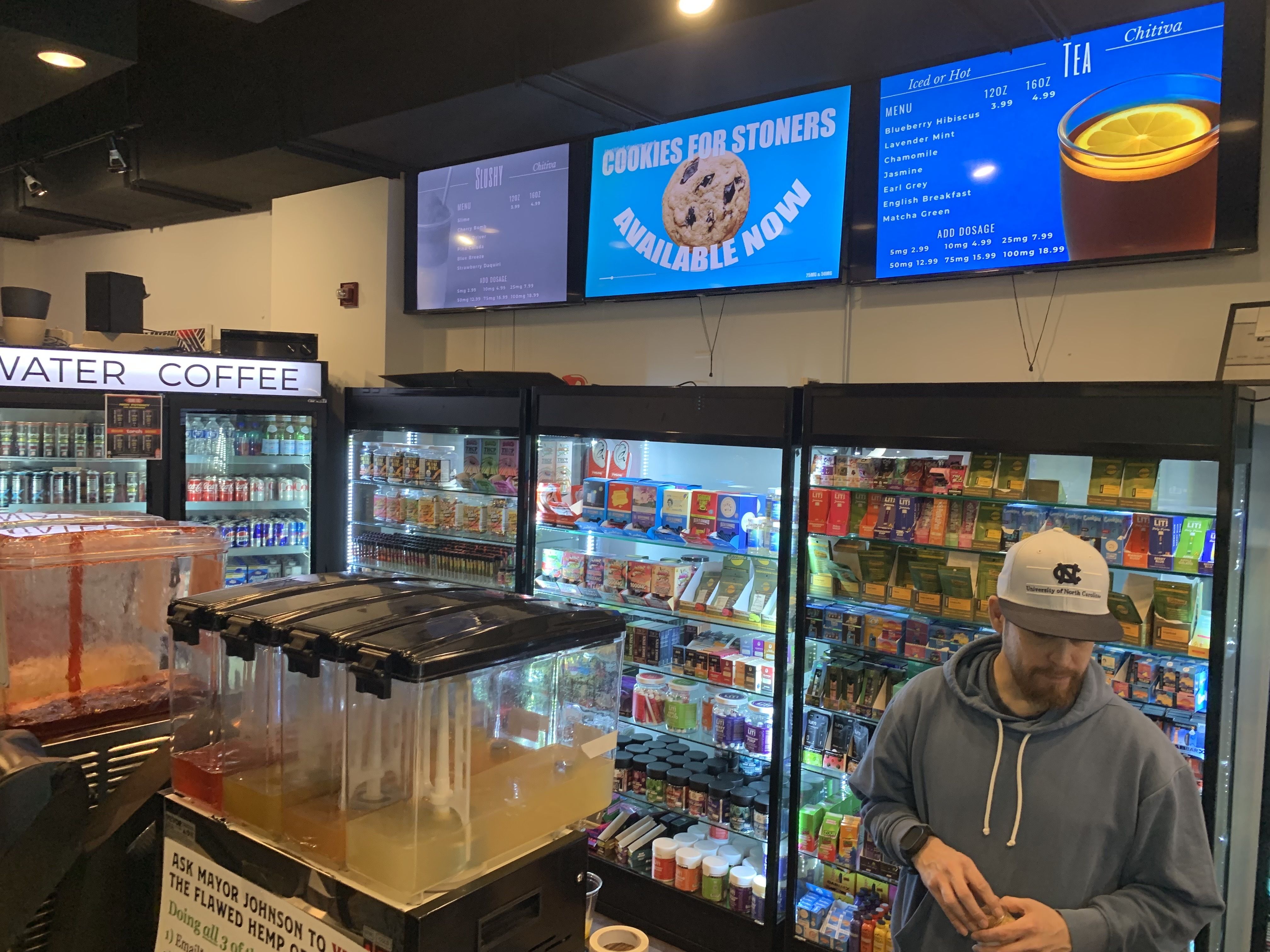 Man wearing a University of North Carolina cap and blue hoodie stands in front of refrigerated shelves stocked with various products in a store with digital menu boards above and drink dispensers in front.