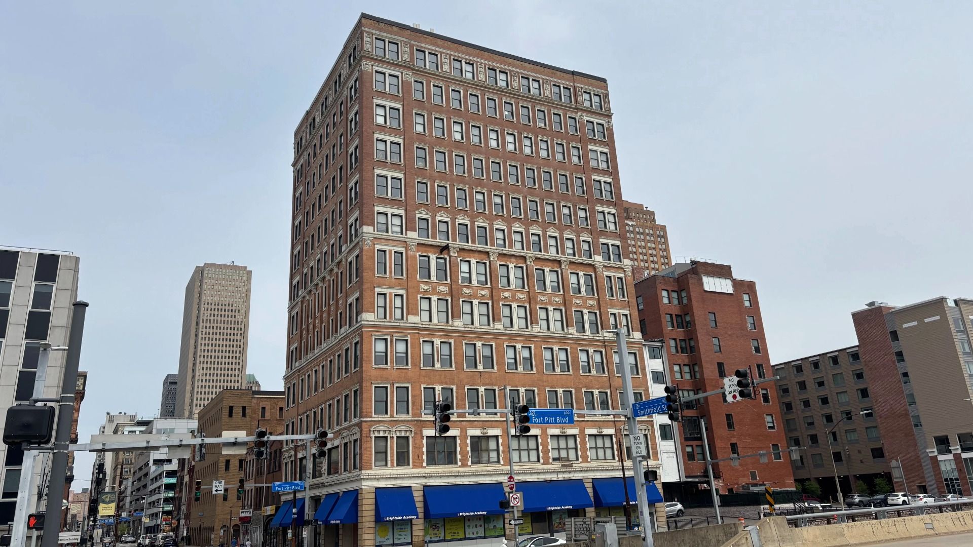 Tall red-brick office building with rows of windows dominates a downtown street; blue storefront awnings line the ground floor, with nearby buildings and a clear blue sky.