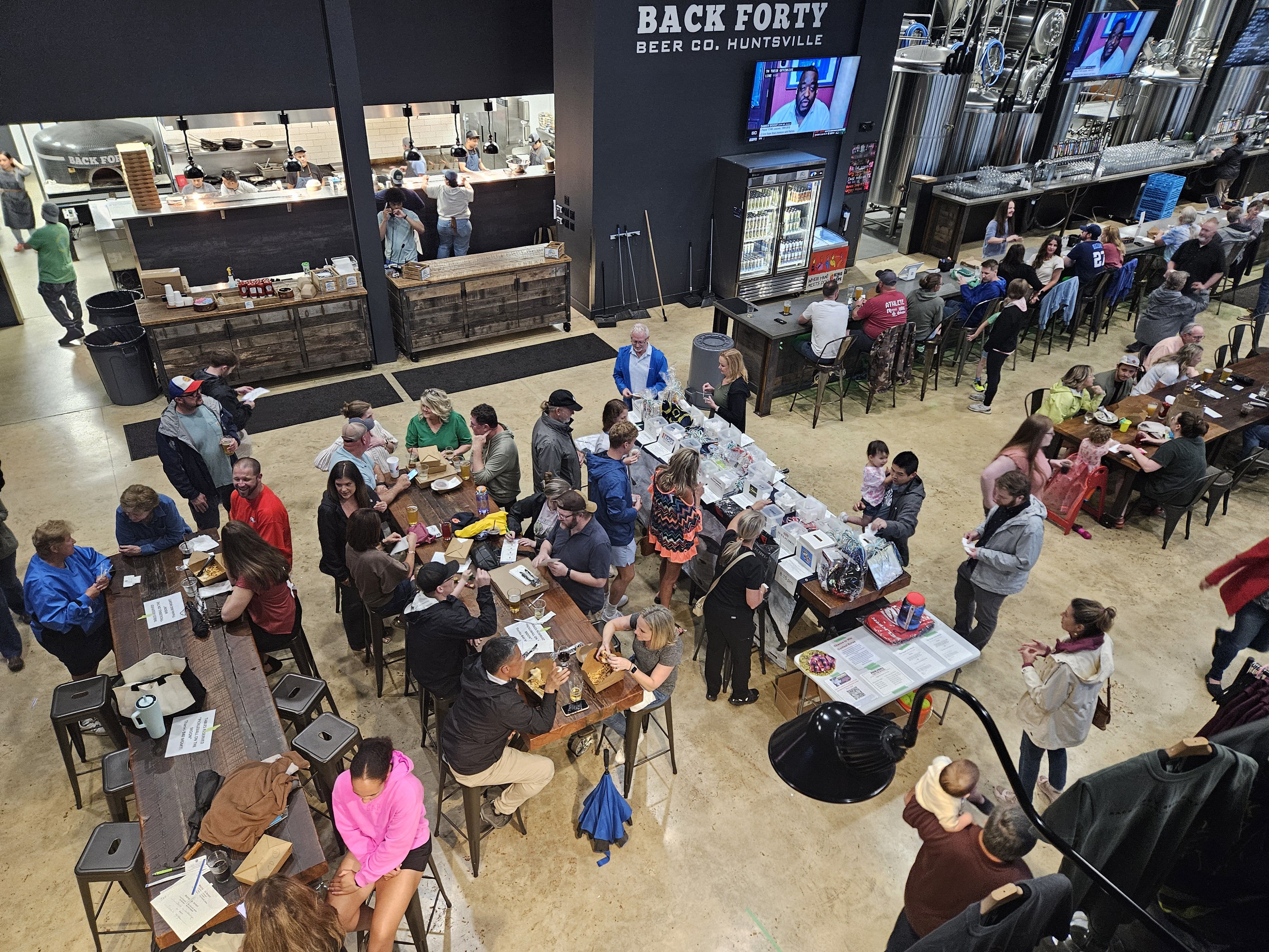Busy Back Forty Beer Co. Huntsville brewery event: long wooden tables filled with people eating, drinking, and chatting, kitchen staff behind a counter, beer taps, refrigerators, and TVs on walls.