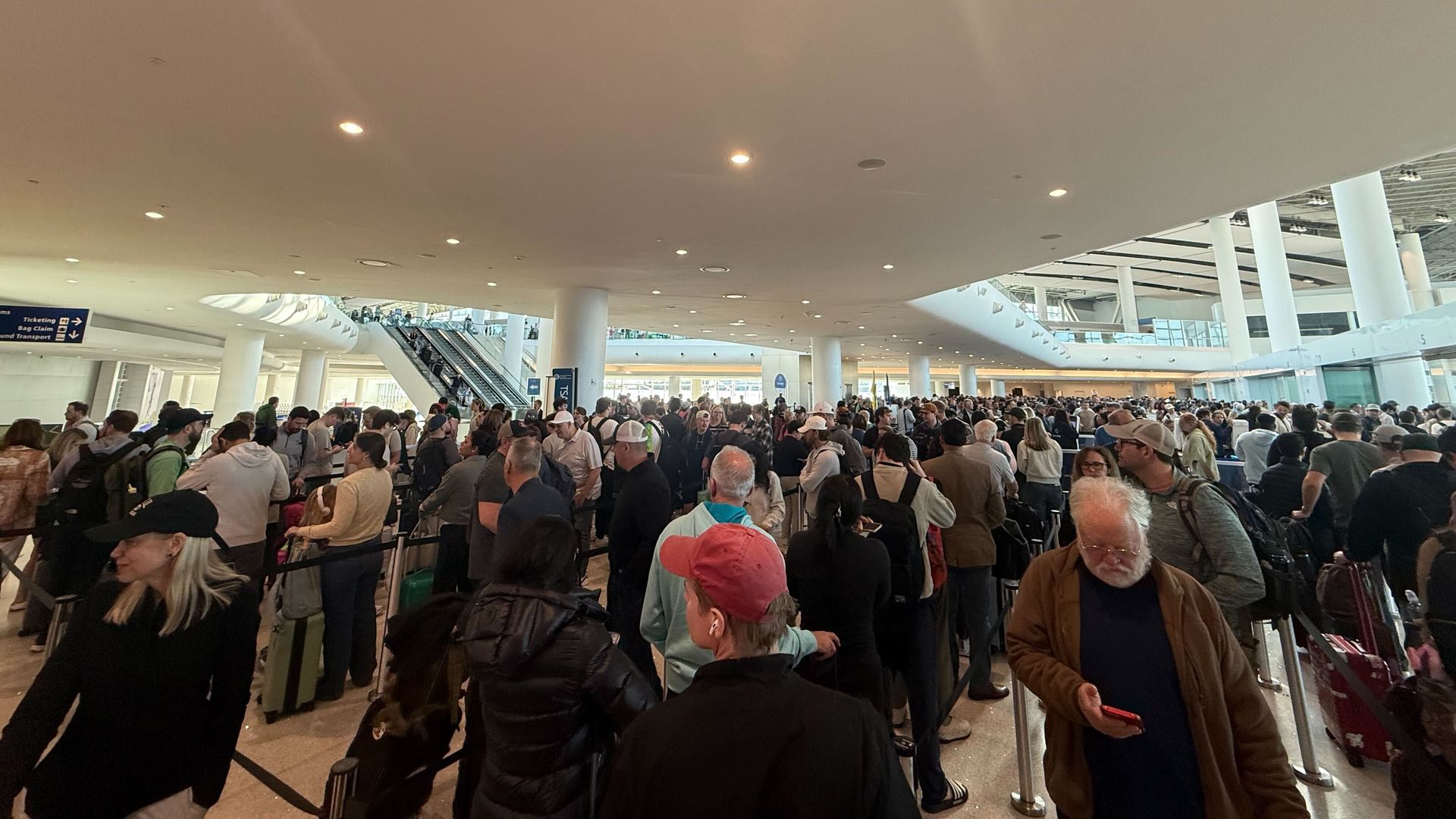 A crowded airport terminal with long lines of travelers waiting with suitcases and backpacks. Bright ceiling, pillars, and an escalator in the background.