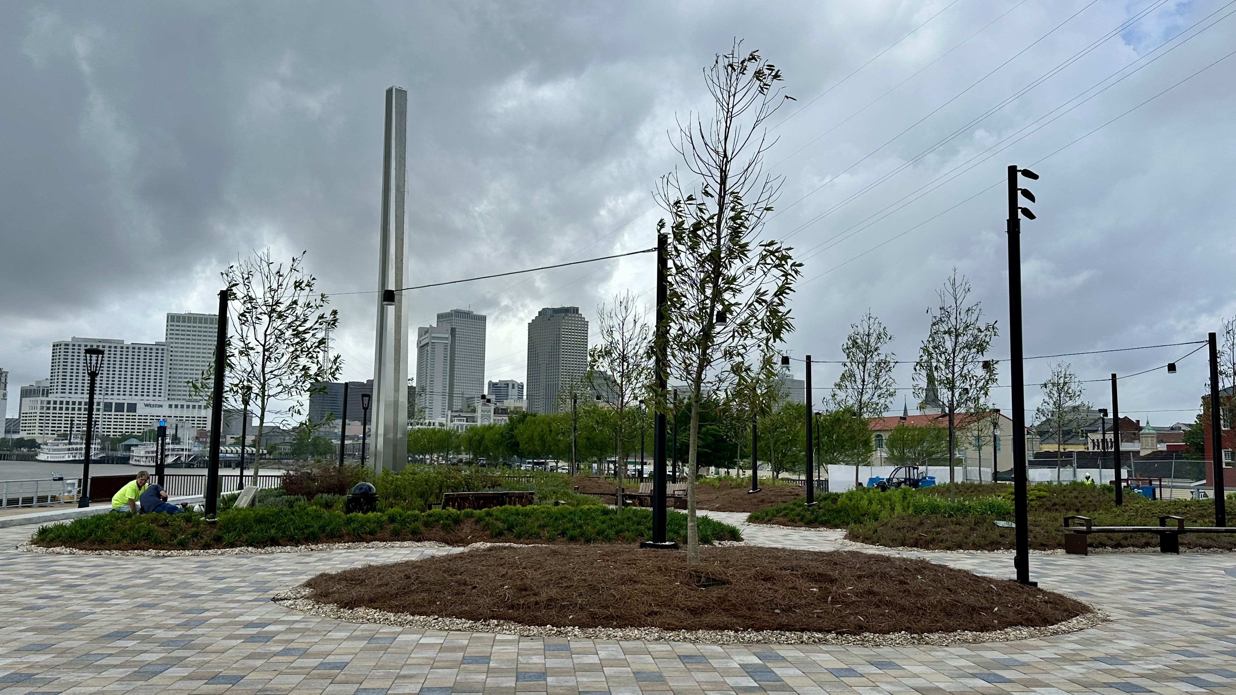 Riverside urban park with a circular mulch island, young trees, and black lampposts on a tiled plaza. A skyline of tall gray buildings under stormy clouds; a pair in neon shirts sits near benches.