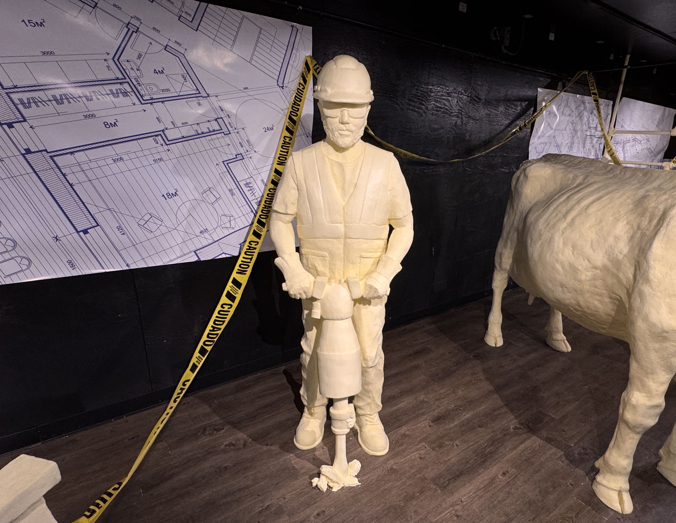 Butter sculpture of a construction worker with a jackhammer, next to a large cow sculpture and blueprints on a black wall, with yellow caution tape in the background.