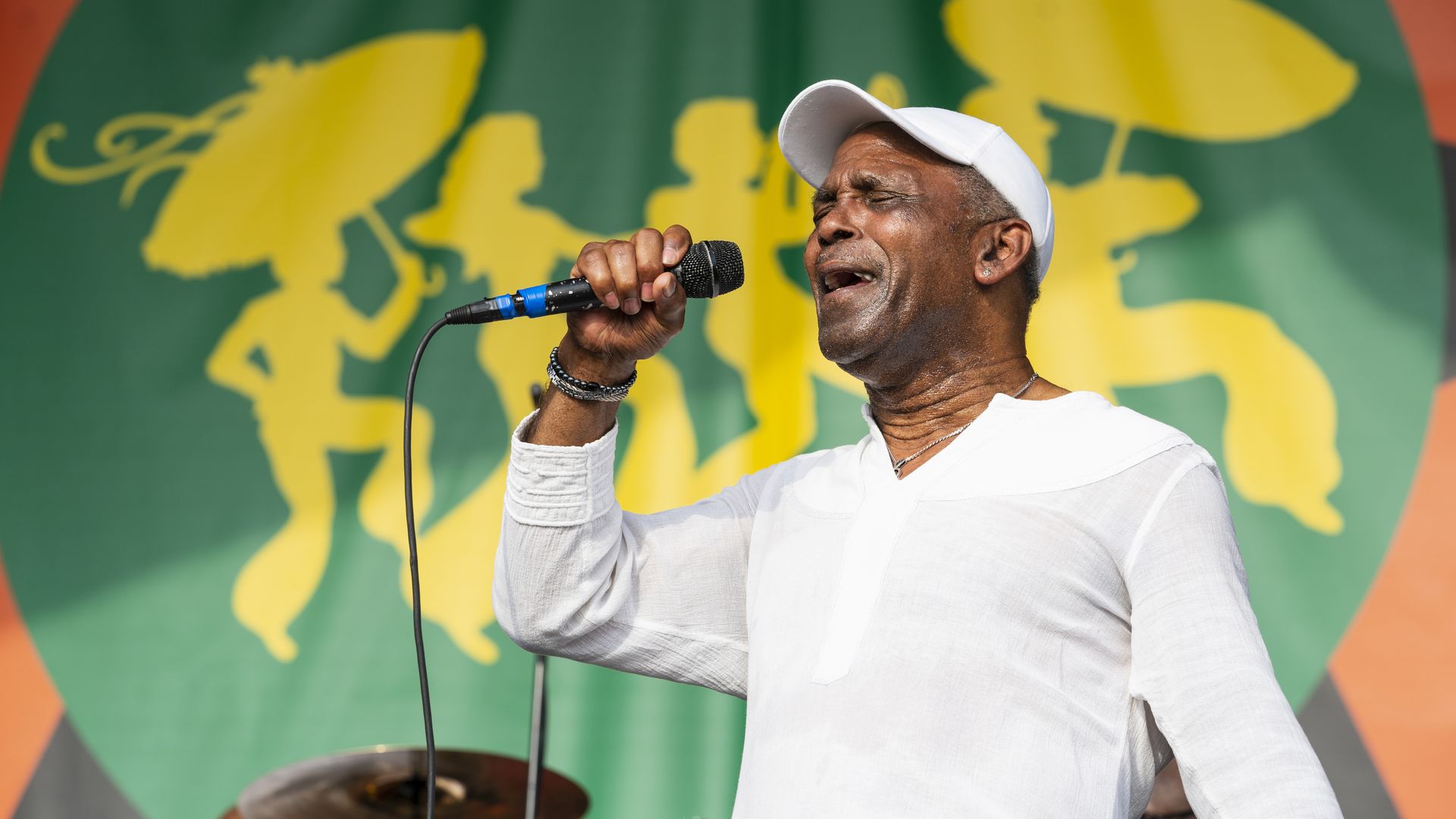 Frankie Beverly sings into a mic on stage at New Orleans Jazz Festival, wearing a white T-shirt and a white hat.