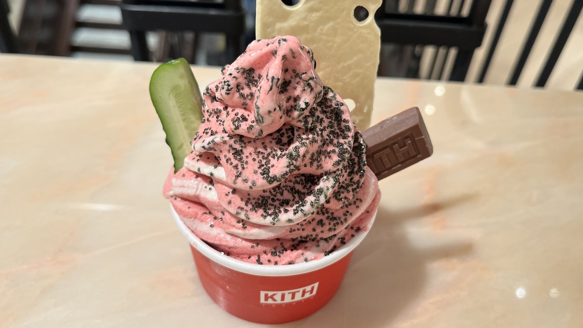 Red KITH cup holds pink swirled soft-serve with black sprinkles, topped by a beige cheese-shaped wafer and a cucumber slice, plus a chocolate bar; light tabletop and striped chairs in the background.