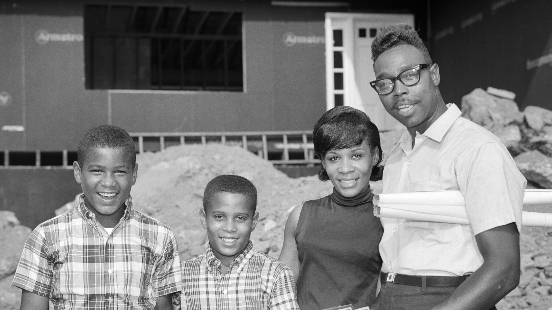 1960s African American family stands before their new home under construction while father holds plans. 