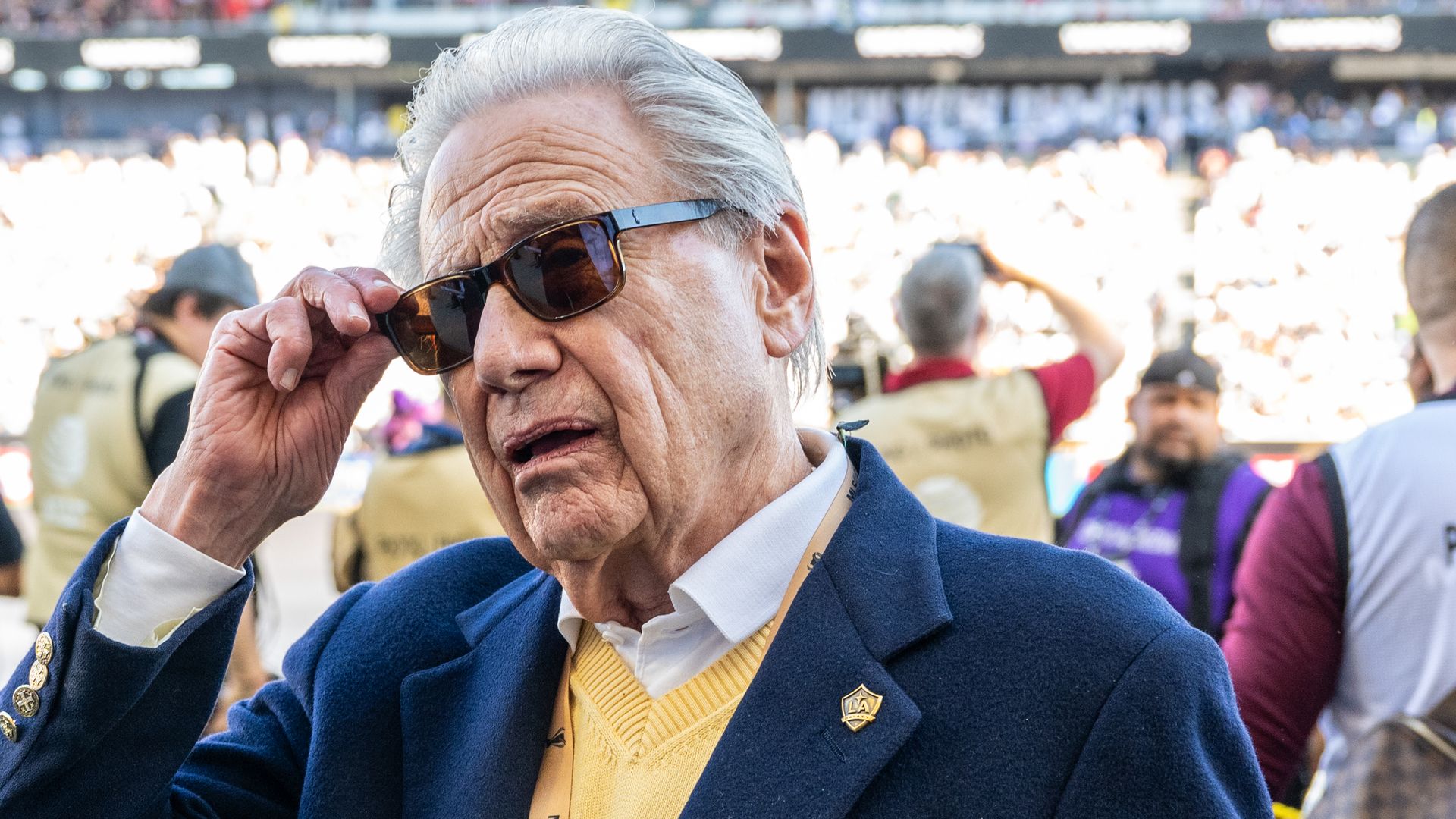 Close-up of an older man with silver hair and dark sunglasses, wearing a blue blazer and yellow sweater, adjusting his glasses at a crowded stadium.