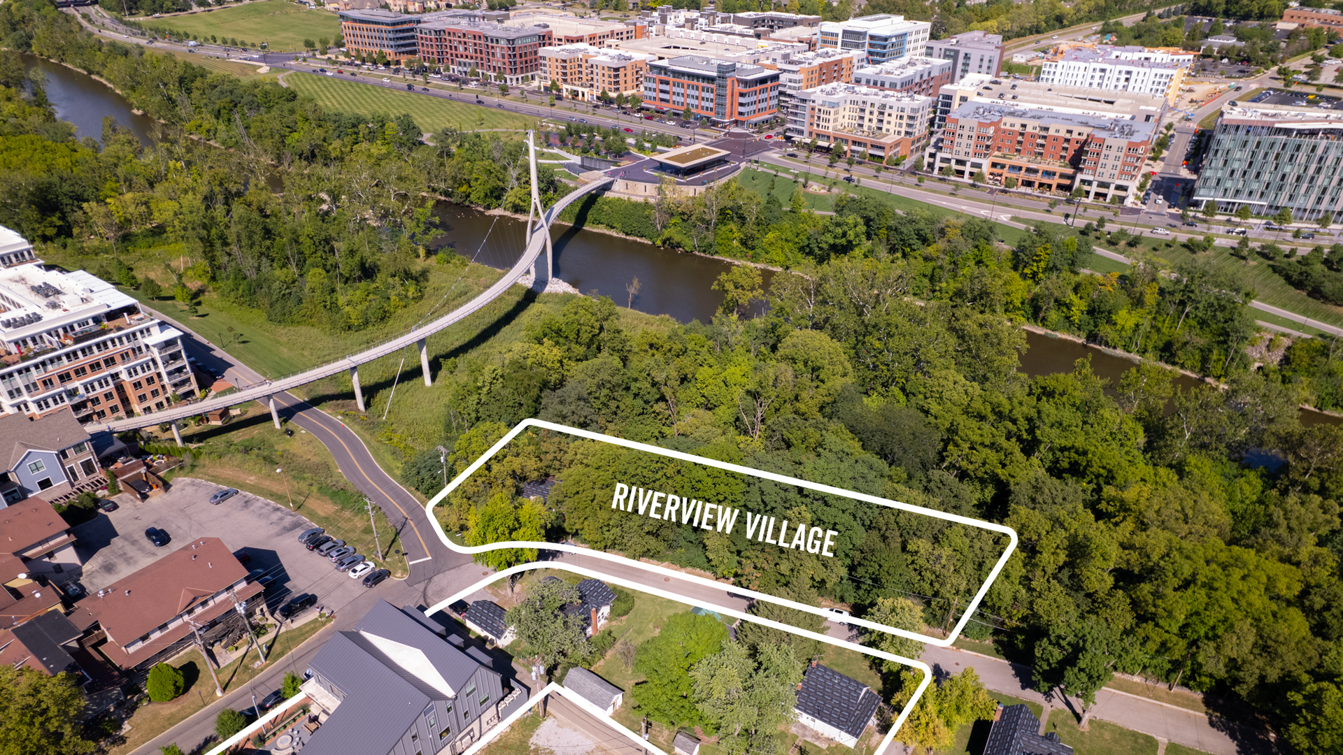 Aerial view of a green wooded area labeled "Riverview Village" near a river with a modern curved pedestrian bridge and clustered buildings in the background under clear skies.