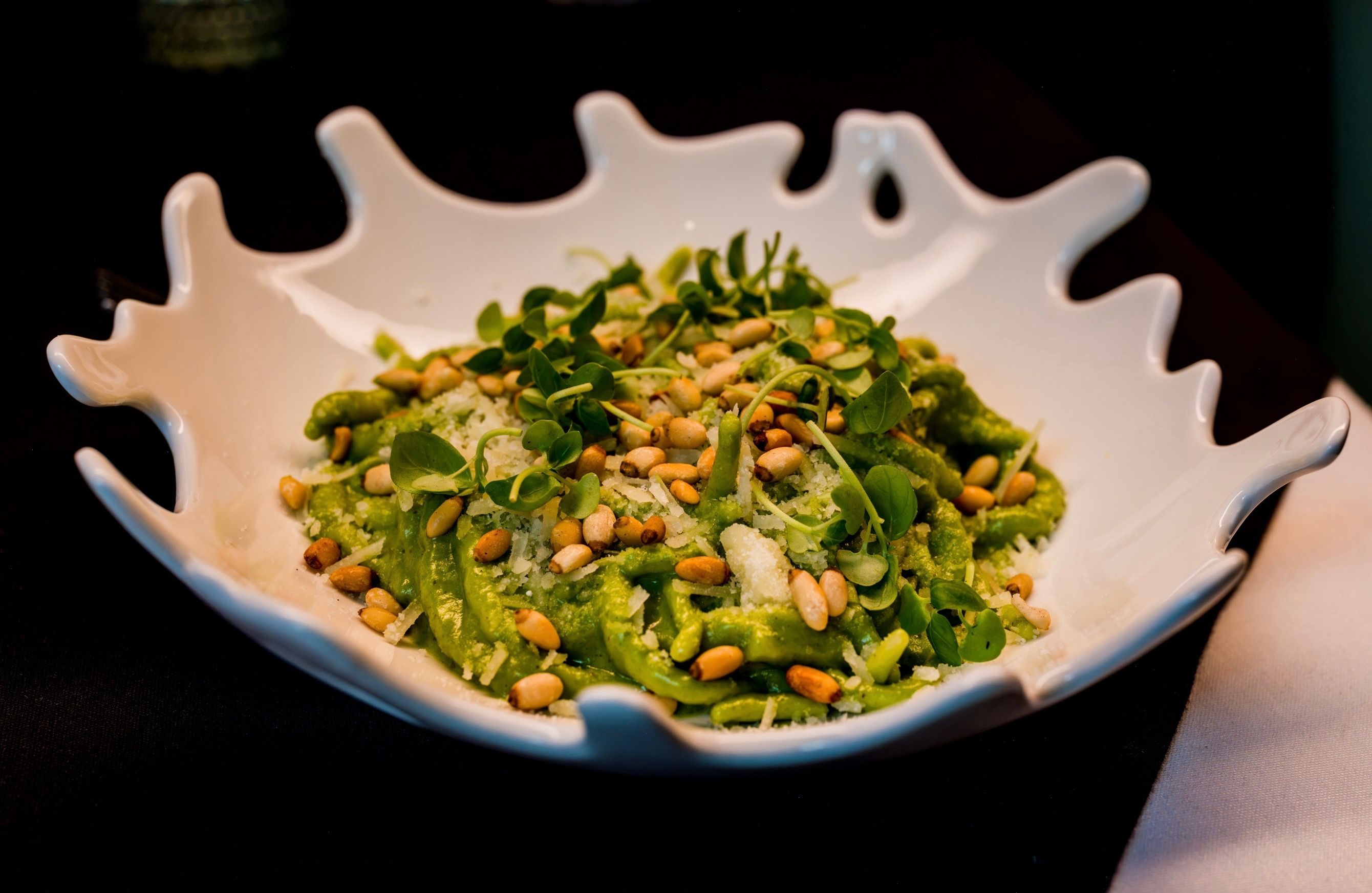 White ceramic bowl with a jagged, wavy rim holds bright green pesto pasta topped with pine nuts and microgreens against a dark background.