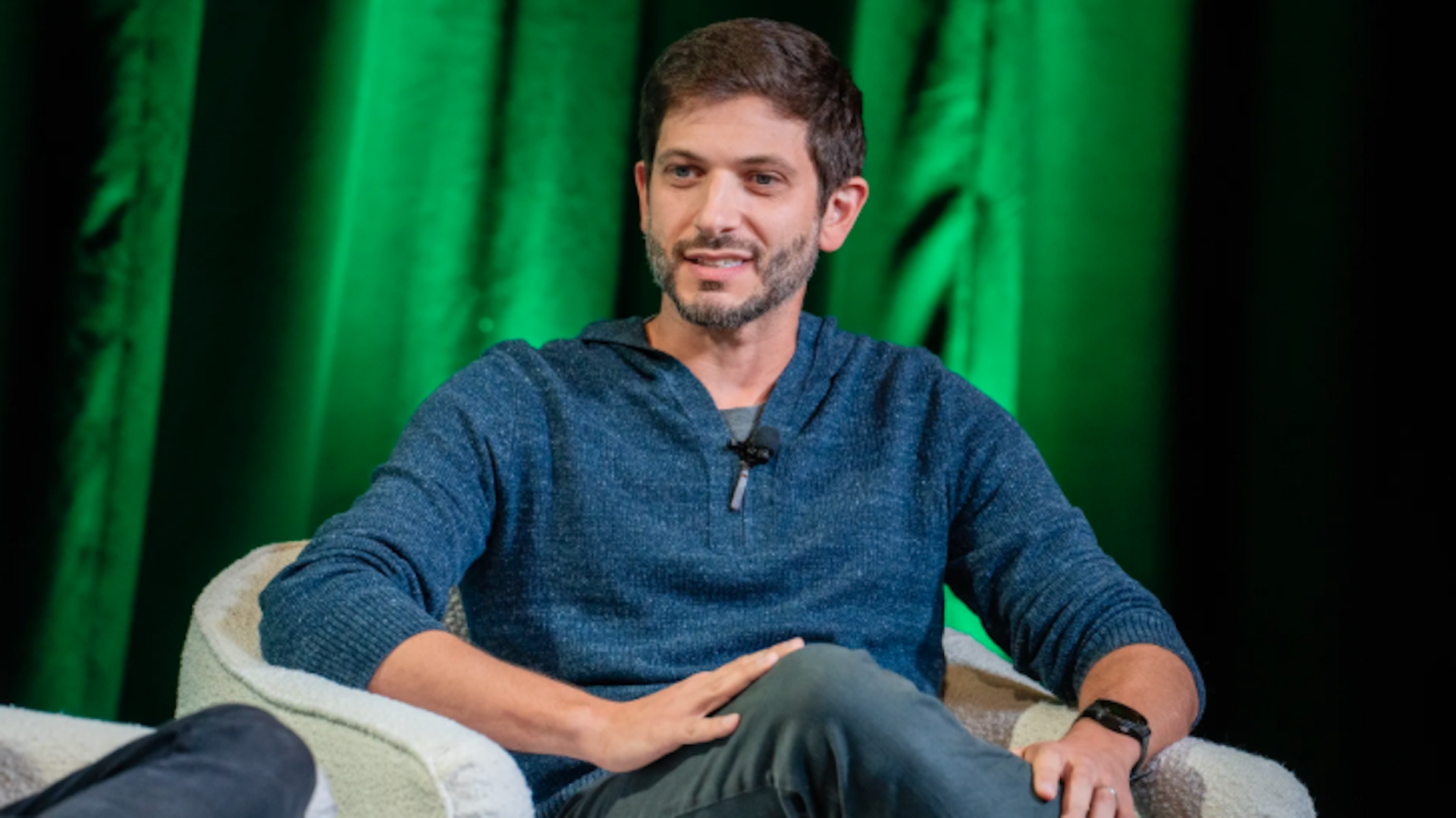 Man with short dark hair and beard sits in a beige armchair on a stage. He wears a blue speckled hoodie and a microphone clipped to his chest; green stage curtains form the background.
