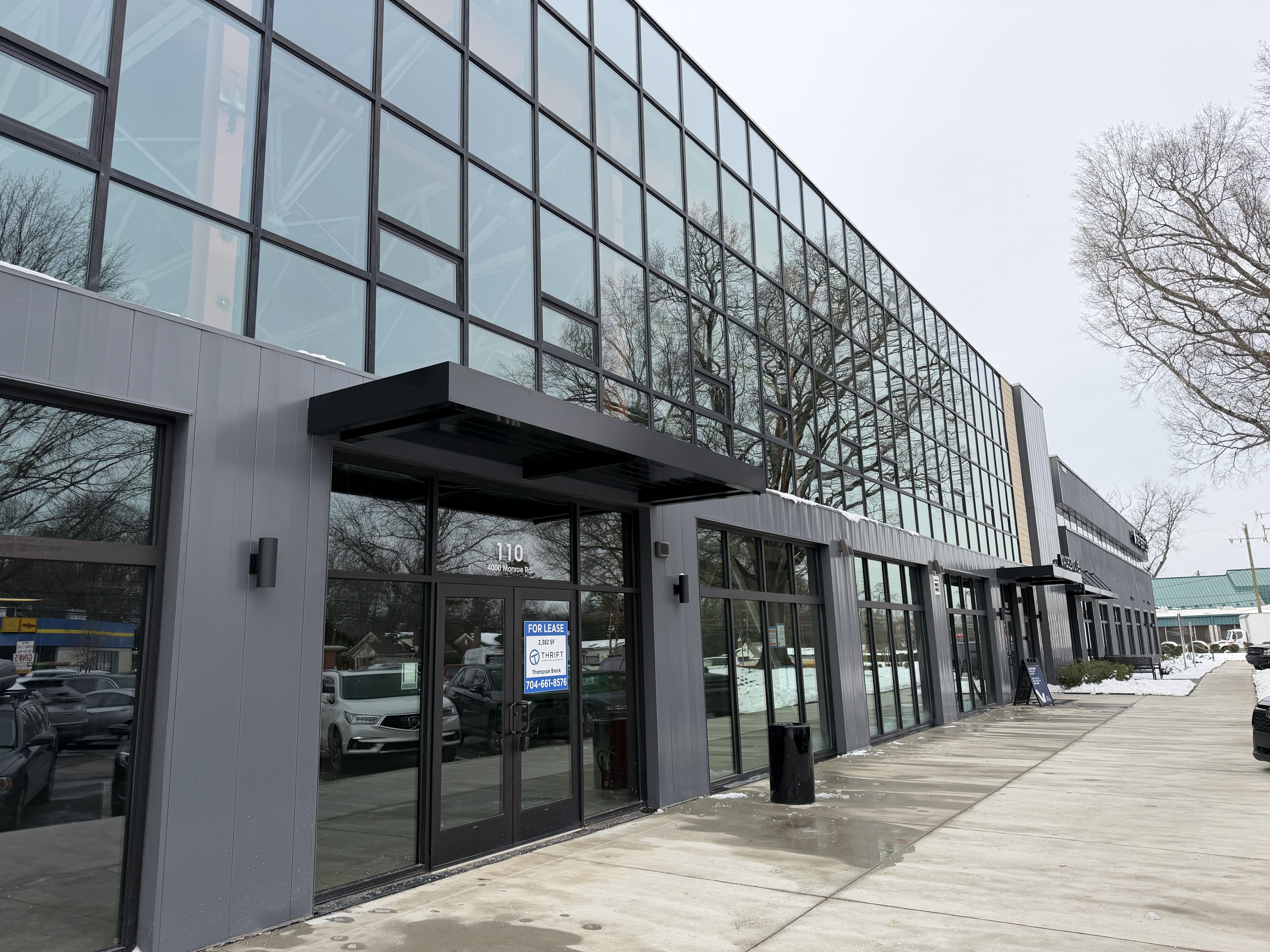 Modern gray commercial building with large glass windows reflecting leafless trees, a concrete sidewalk, a 'For Lease' sign on door, and a cloudy winter sky.