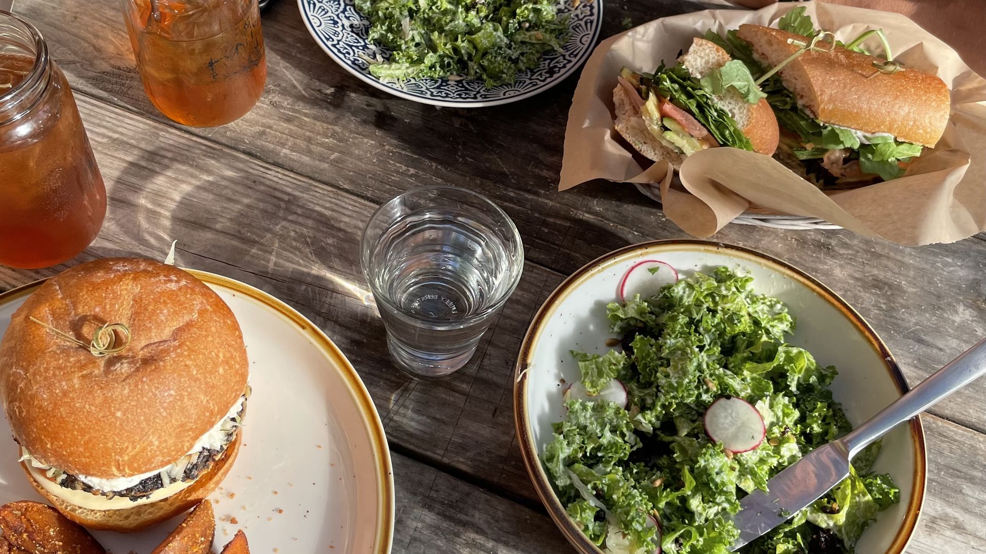 Table with a burger on a white plate, wedge-cut fries, two glasses of iced tea, a sandwich with greens on parchment, and two bowls of kale salad with radish slices and dressing.